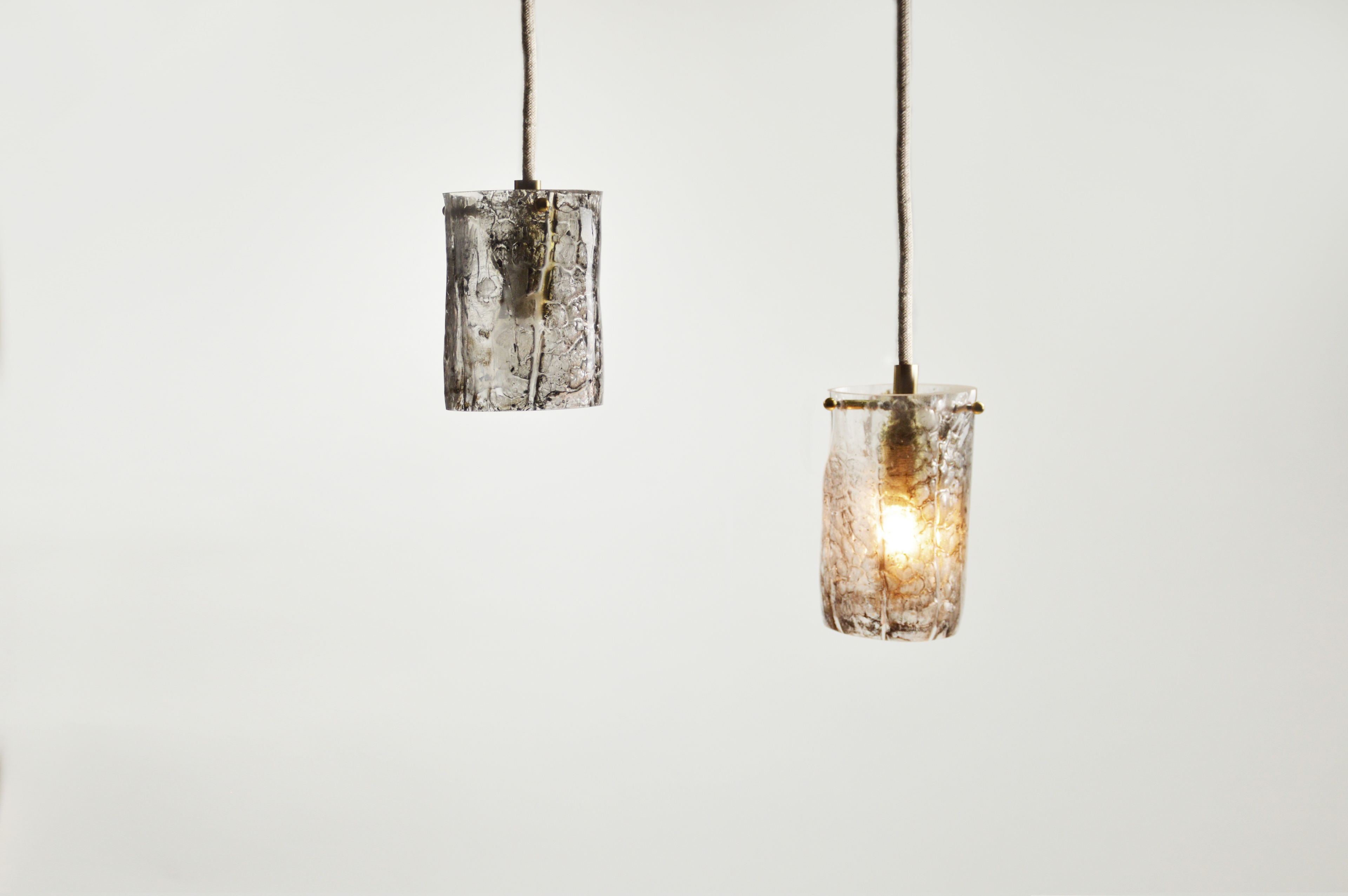 Suspension lumineuse en verre au décor interne d’écorce de bois, obtenu par prise d’empreinte d’une branche, enrobée de verre en fusion. Verre teinté naturellement par le processus de mise en forme, dépoli et voiles noirs de combustion, aléatoires et uniques.