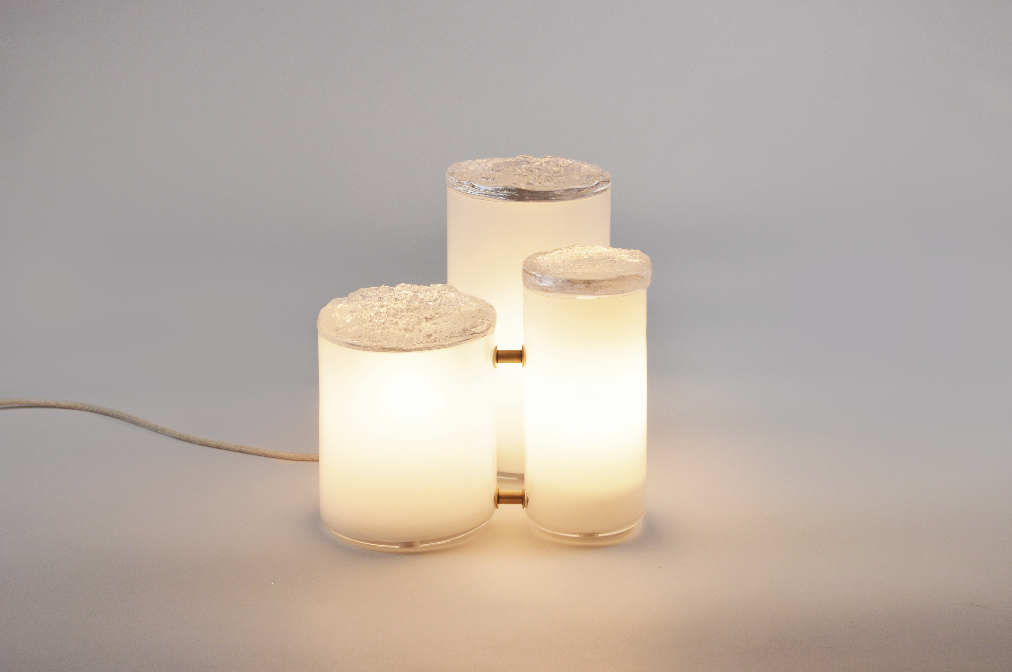 Luminaire composé de trois cylindres de verre soufflé opalin, surmontés de couvercles de verre massif. Empreintes de bois obtenues par trois coulées de verre en fusion sur une même souche. Trois formes réunies en fagot, détails de laiton. h : 310 mm L : 320 mm l : 280 mm