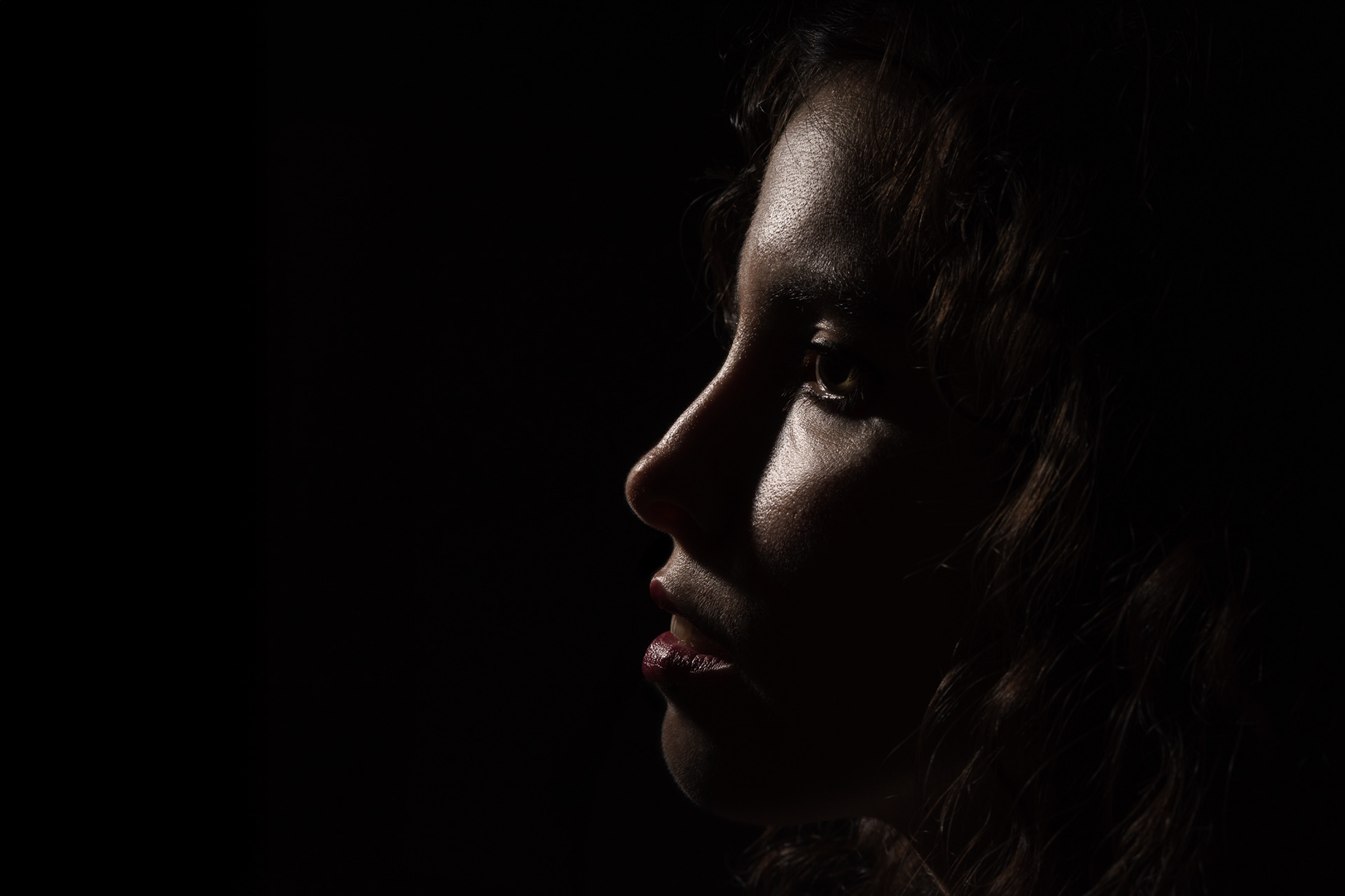 Retrato low key de mulher de perfil com luz dura realçando os contornos, iluminação dramática estilo Rembrandt sobre fundo preto.