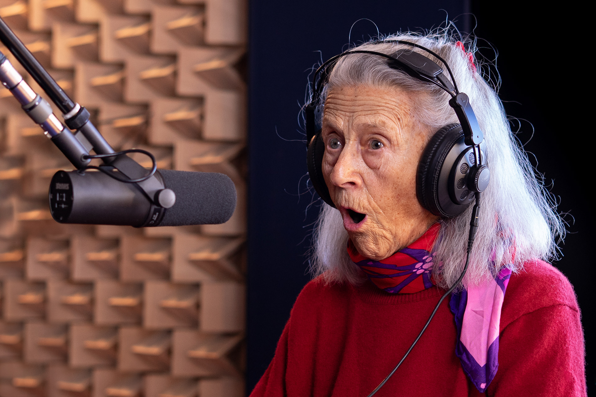 Retrato muito expressivo de mulher idosa de olhos claros, com lenço ao pescoço e camisola vermelha, com headphones na cabeça, atrás de um microfone Shure SM7B, num estúdio de som em Linda-a-Velha, junto a Lisboa, durante gravação e performance musical. A mulher está com a boca aberta a cantar ou em performance vocal e percebe-se que está a interagir com alguém fora do enquadramento. Ao fundo, desfocados, vêem-se esponjas acústicas. A fotografia foi iluminada com luzes contínuas e softboxes.