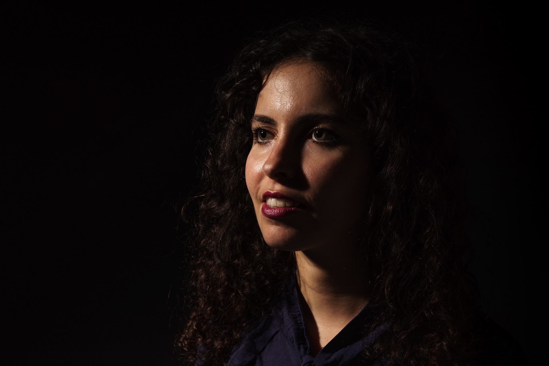 Retrato de mulher jovem com rosto e pescoço parcialmente iluminados por luz dura, inspirado na iluminação Rembrandt, fundo preto, fotografia de estúdio em Lisboa.