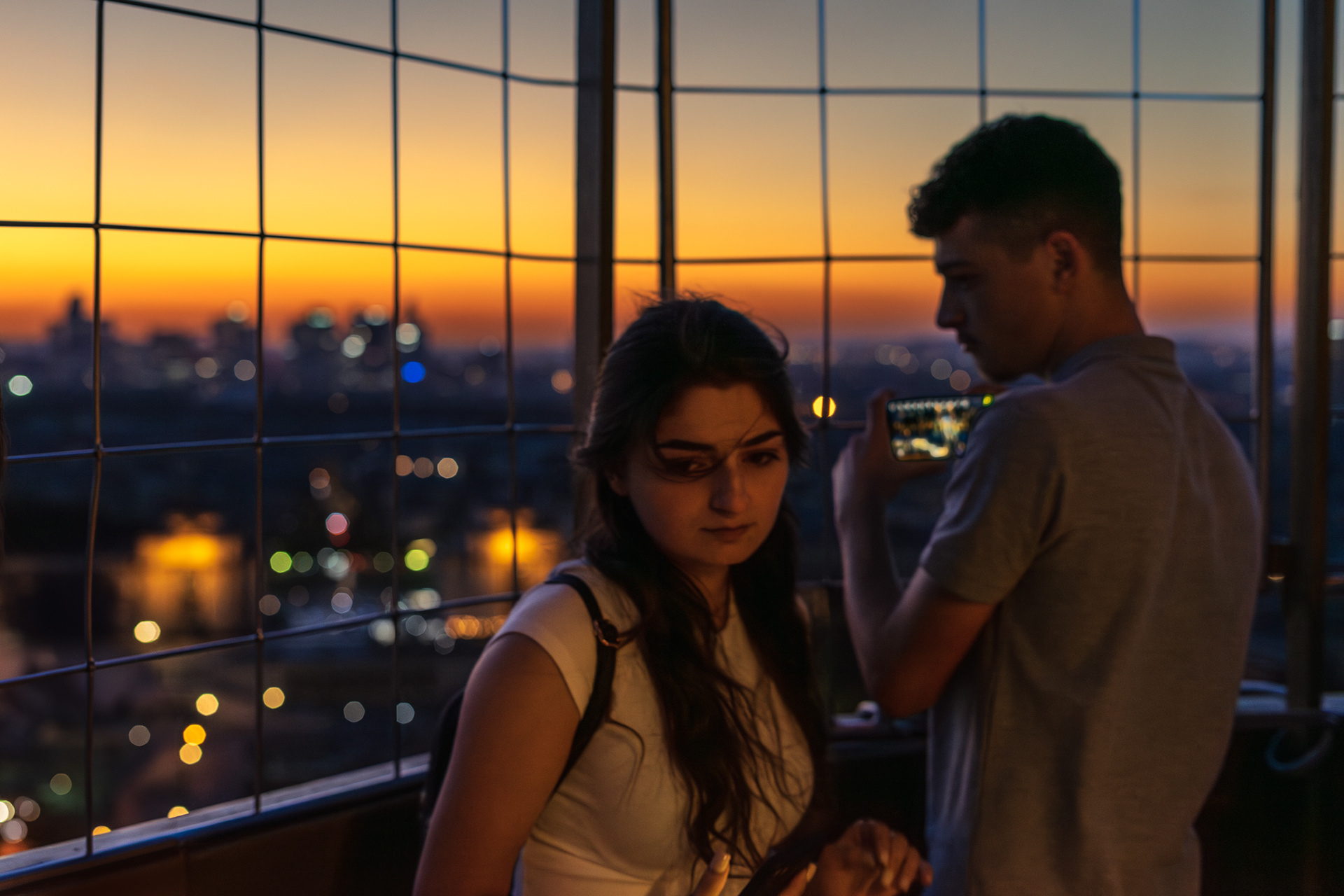 Retrato de mulher jovem de frente a olhar para baixo com expressão ligeiramente triste, acompanhada por homem jovem atrás meio de perfil também a olhar para baixo, fotografia no interior da Torre Eiffel em Paris ao final do dia com luz dourada, paisagem urbana desfocada e luzes desfocadas ao fundo.