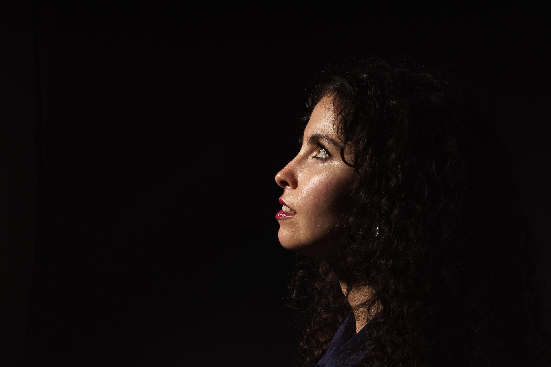 Retrato low key de mulher jovem de perfil a olhar ligeiramente para cima, com iluminação dura destacando a face bem iluminada, cabelo parcialmente na sombra, fundo preto, fotografia de estúdio em Lisboa.