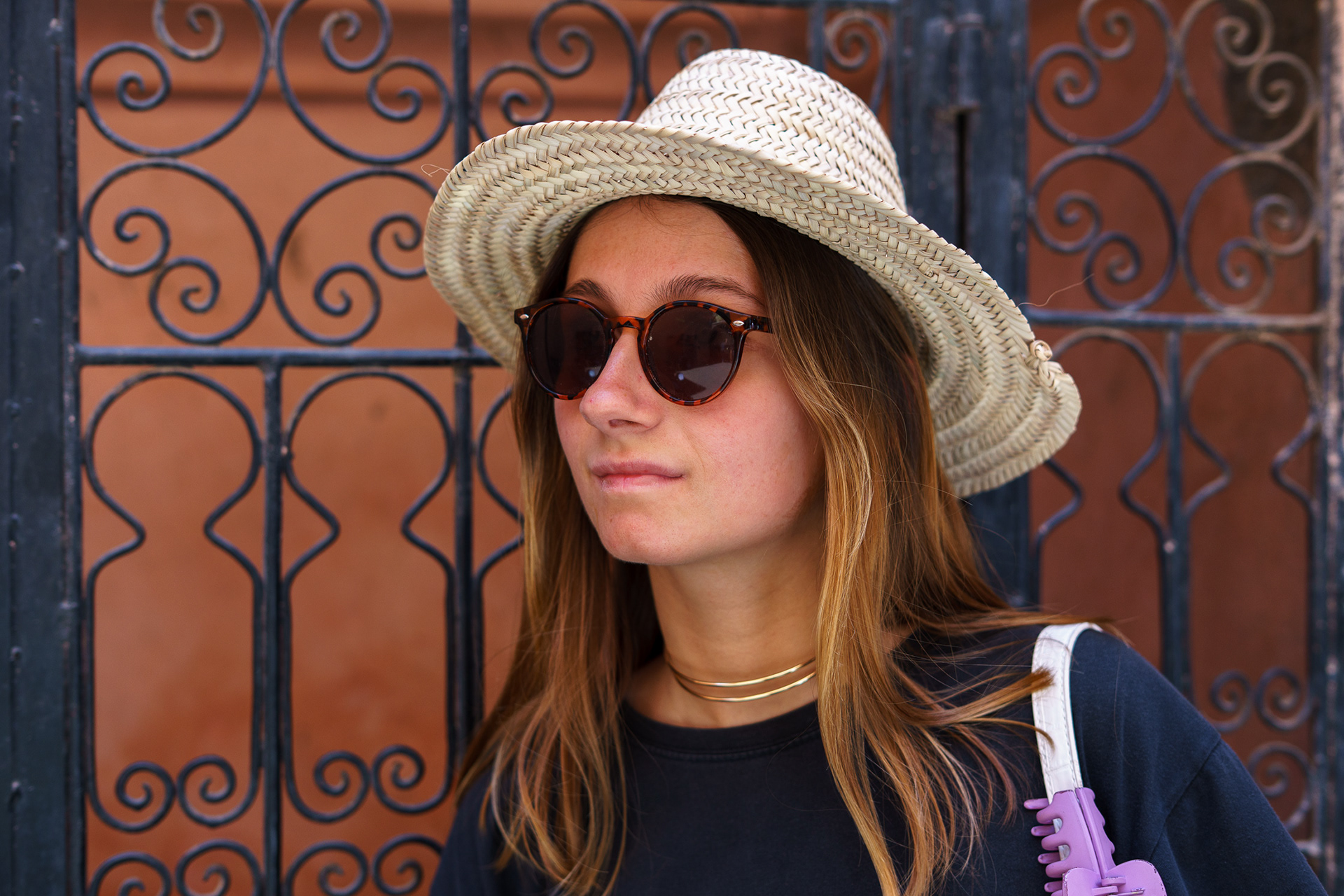 Retrato de mulher muito jovem de chapéu claro e óculos escuros a olhar ligeiramente para o lado, tirado na rua em Marraquexe, Marrocos, com portão e parede de cor quente e vibrante ao fundo, iluminação natural.