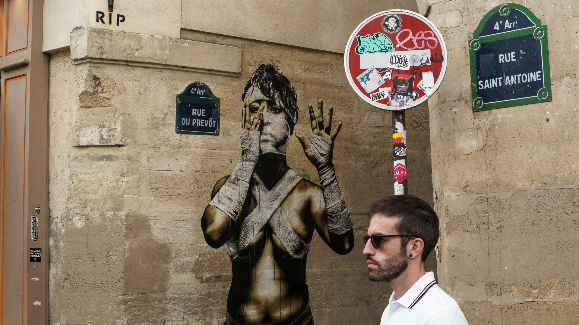 Retrato urbano em Paris de homem de perfil com camisola branca e óculos escuros, luz natural, junto a parede com pintura de mulher com ligaduras na cara, braços e corpo a tapar o peito, placas de rua em francês e sinal de proibido.