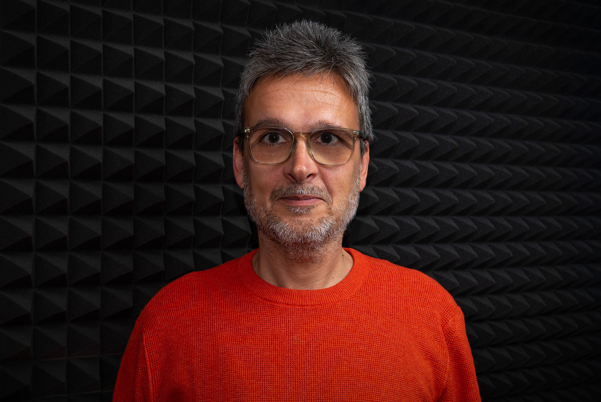 Auto-retrato de Rodolfo Martins, fotógrafo especializado em produto e retrato, a olhar para a câmara, com óculos e expressão semi-sorridente, em frente a esponjas acústicas cinzentas escuras que conferem profundidade e tridimensionalidade ao retrato. Fotografia captada em estúdio com iluminação profissional de três pontos (flash, luz rebatida no teto e luz suave lateral com softbox). Imagem usada na secção ‘Sobre mim’ do site, convidando a conhecer o trabalho e a contactar para novos projetos fotográficos.