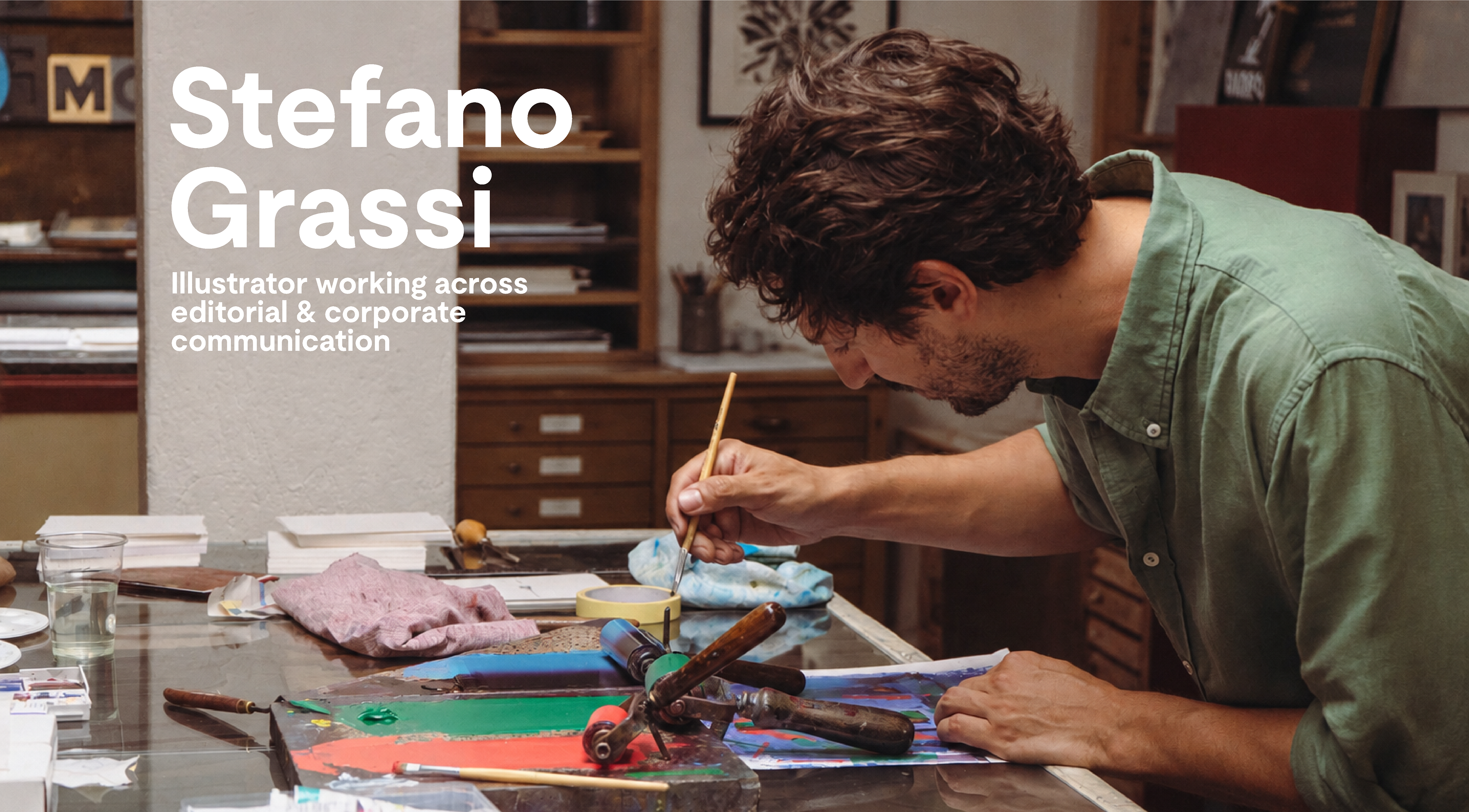 Fotografia di Stefano Grassi mentre lavora nell'atelier. Sul tavolo si vedono inchiostri colorati stesi con spatole e rulli, Asquarelli e tubi di colore. Stefano sta inchiostrando usando un pennello