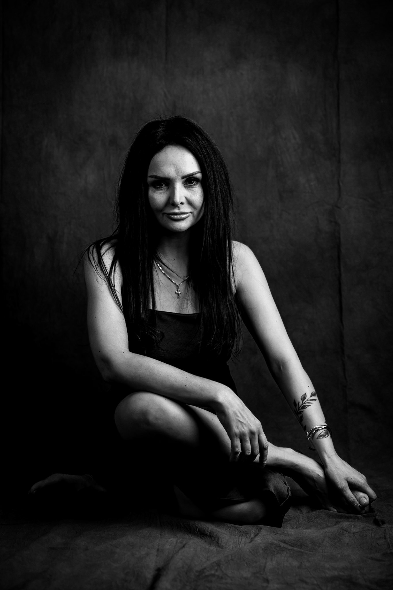 Full-length black-and-white studio portrait of a woman seated against a dark textured backdrop.