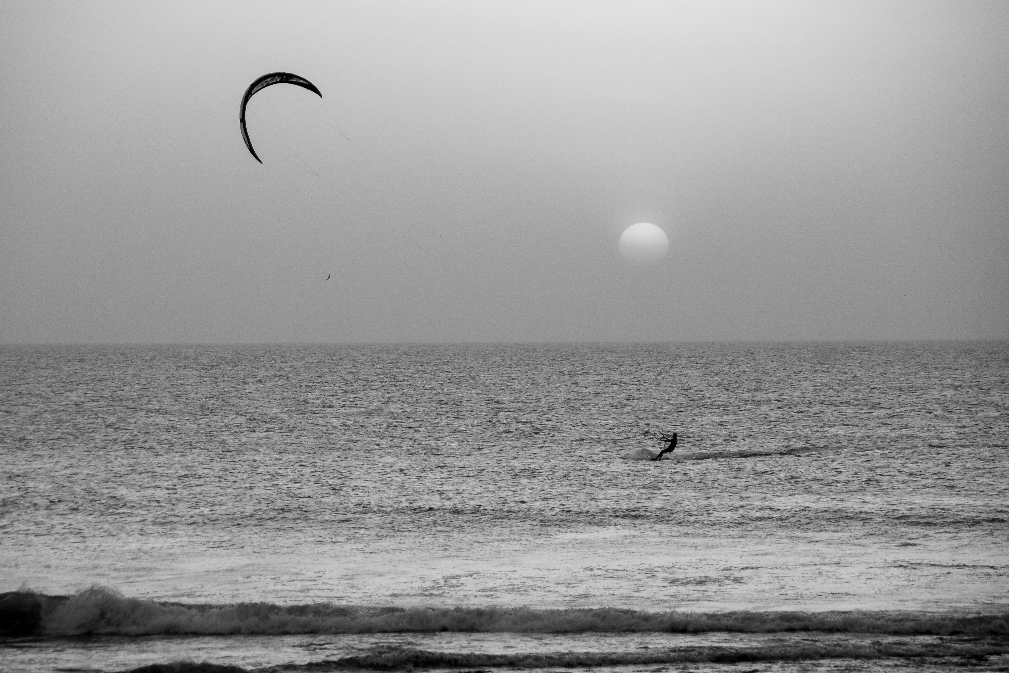 Black-and-white seascape with a lone kitesurfer beneath a hazy sun.