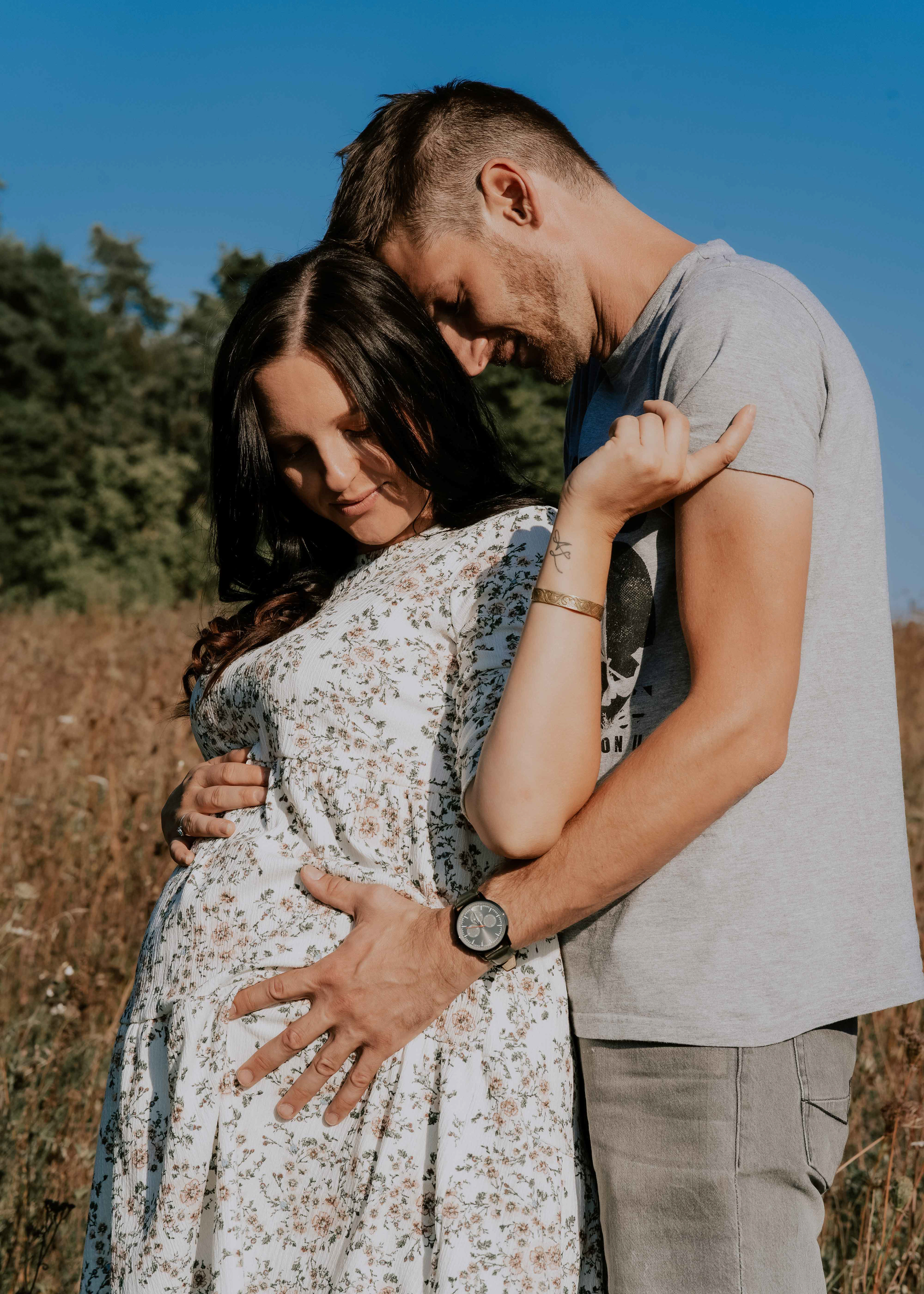 Ein liebevolles Partnerbild eines Paares beim Babybauch-Shooting in Regensburg, bei dem der werdende Vater den Bauch seiner Partnerin im Blumenkleid zärtlich umschließt.