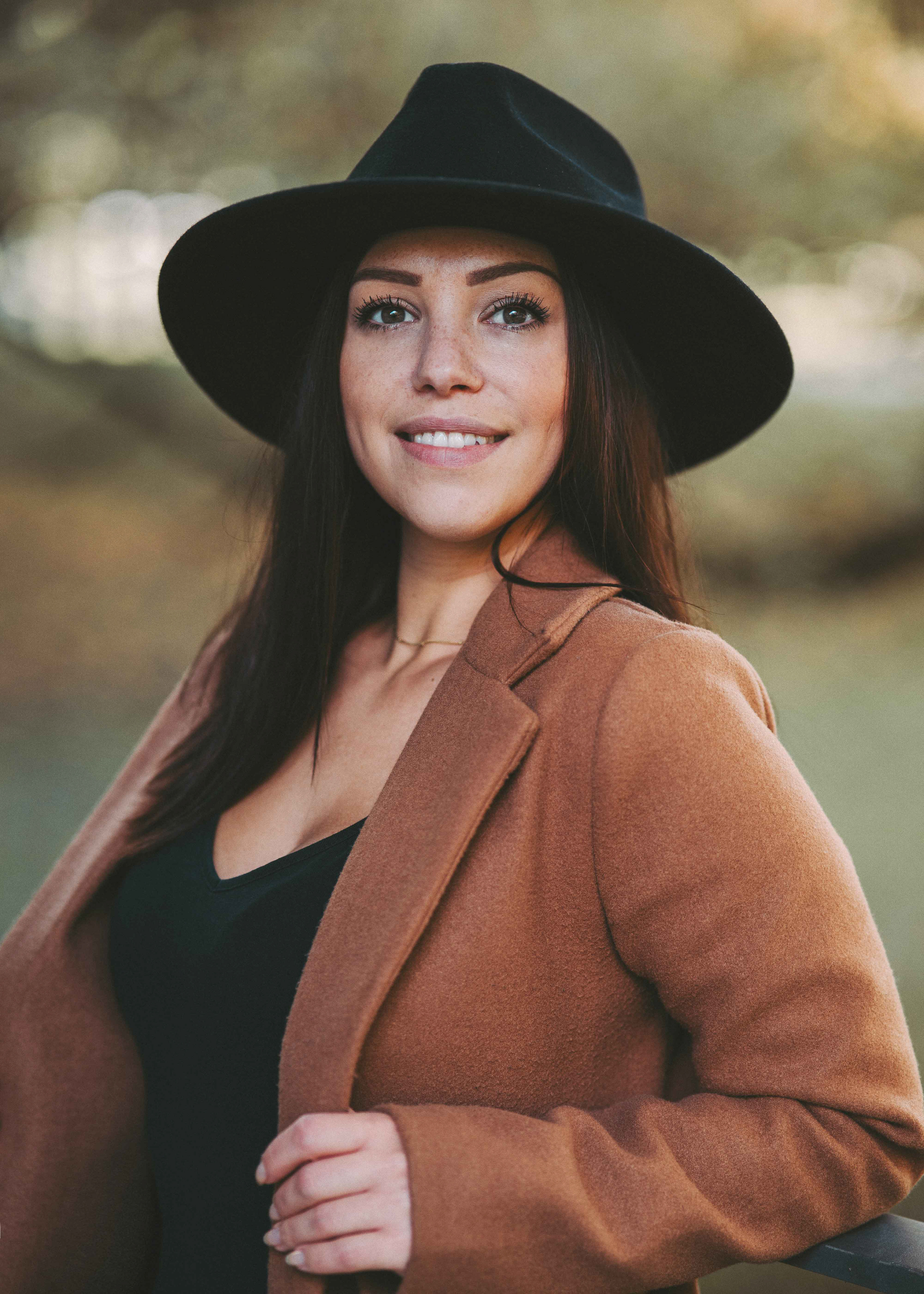 Porträt einer lächelnden Frau im Stadtpark Regensburg, fotografiert von Medusa Fotografie, mit braunem Mantel und schwarzem Hut.