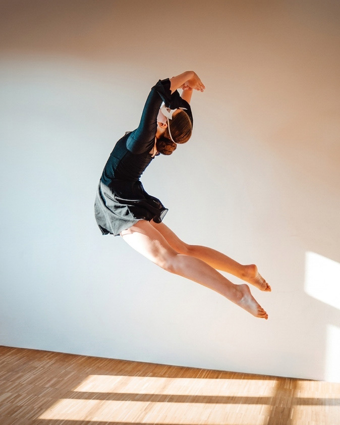 Eine Tänzerin im schwarzen Dress zeigt eine dynamische Sprungpose in der Luft vor einer minimalistischen weißen Wand.