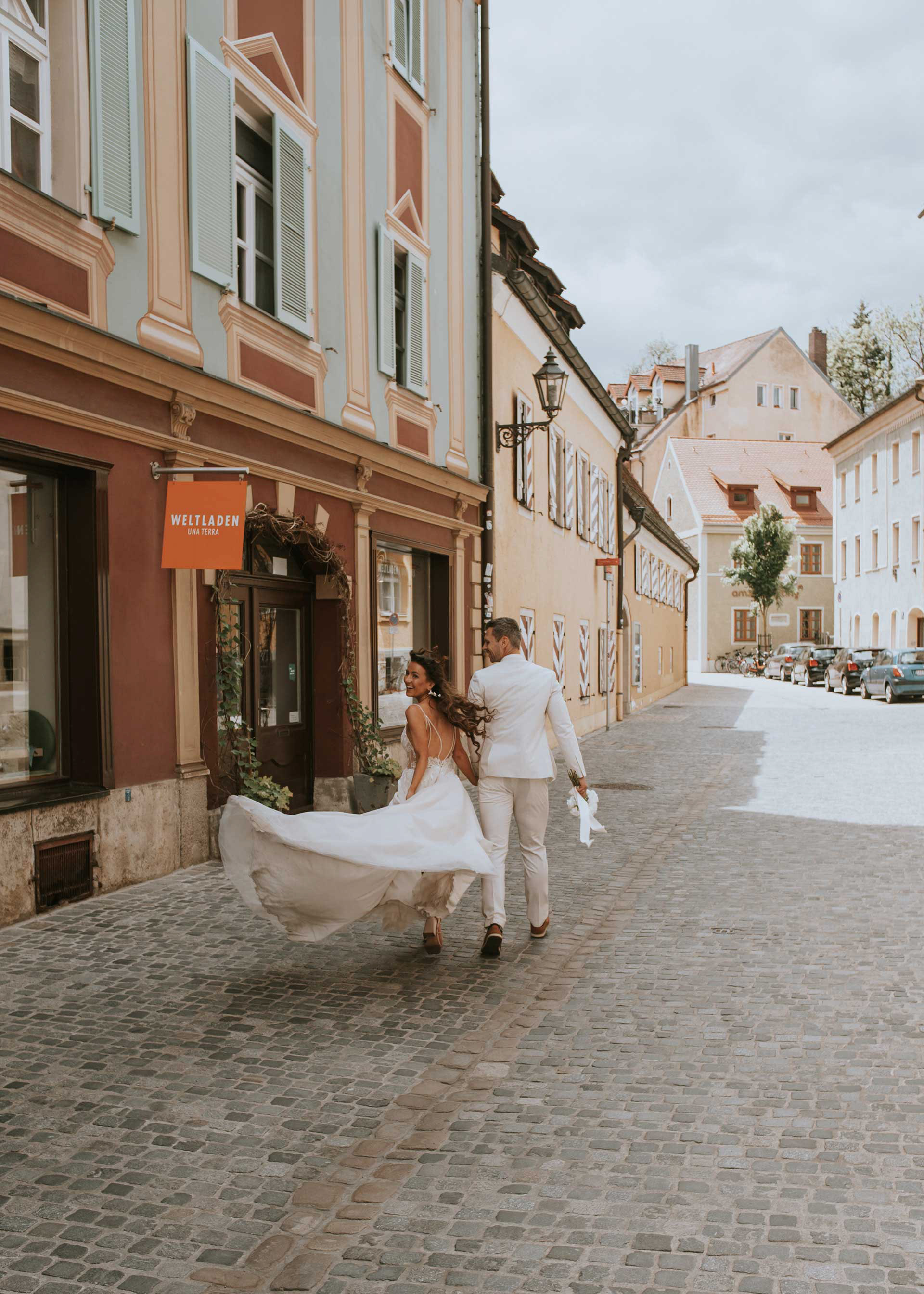 Ein Brautpaar läuft Hand in Hand durch eine gepflasterte Altstadtstraße. Die Braut im bestickten Hochzeitskleid blickt zurück und lächelt. Der Bräutigam im weißen Anzug hält einen Brautstrauß.