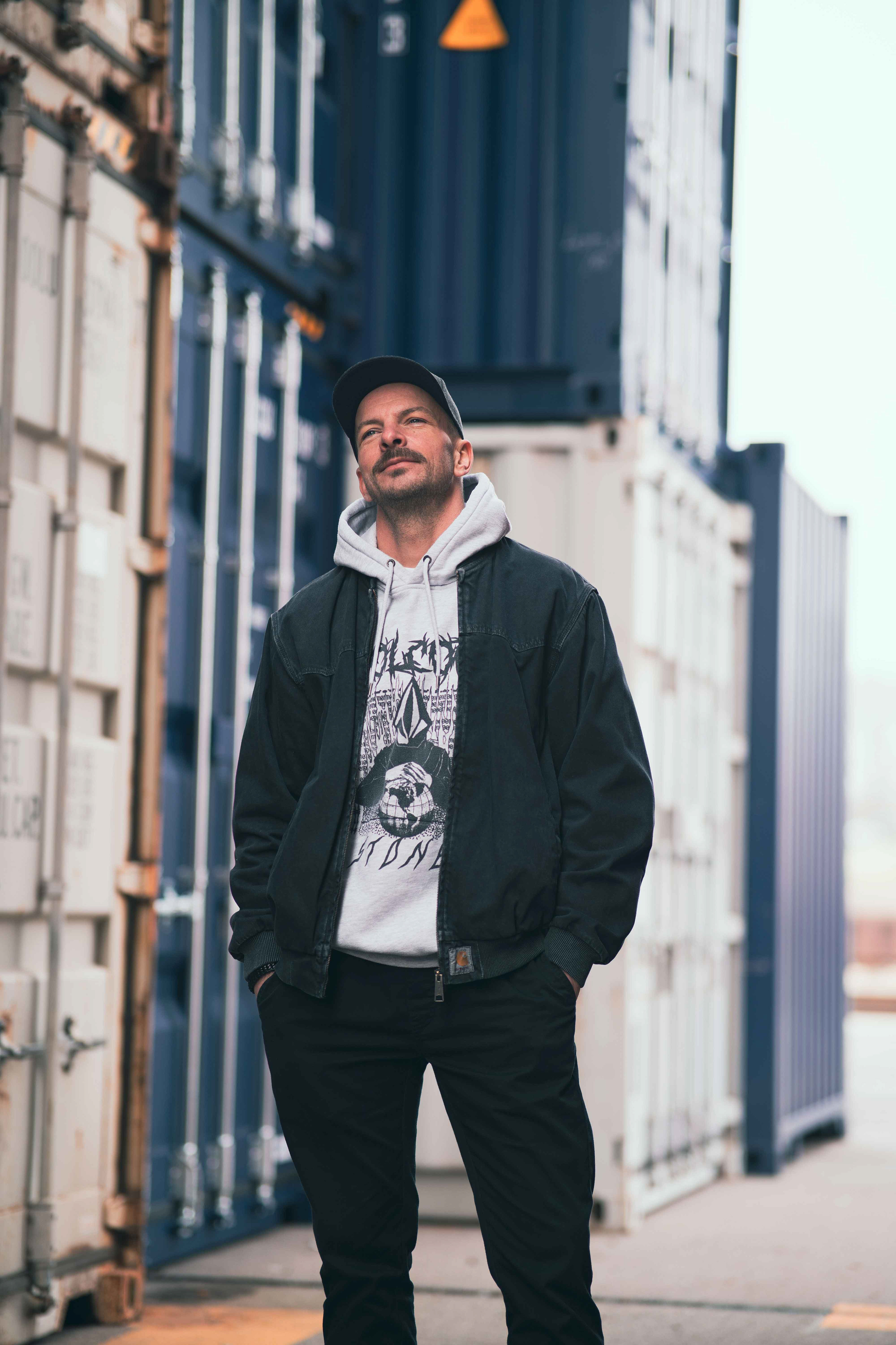 Streetstyle-Foto eines Mannes in schwarzer Jacke und hellem Hoodie vor blauen Schiffscontainern im Regensburger Hafen, fotografiert von Medusa Fotografie