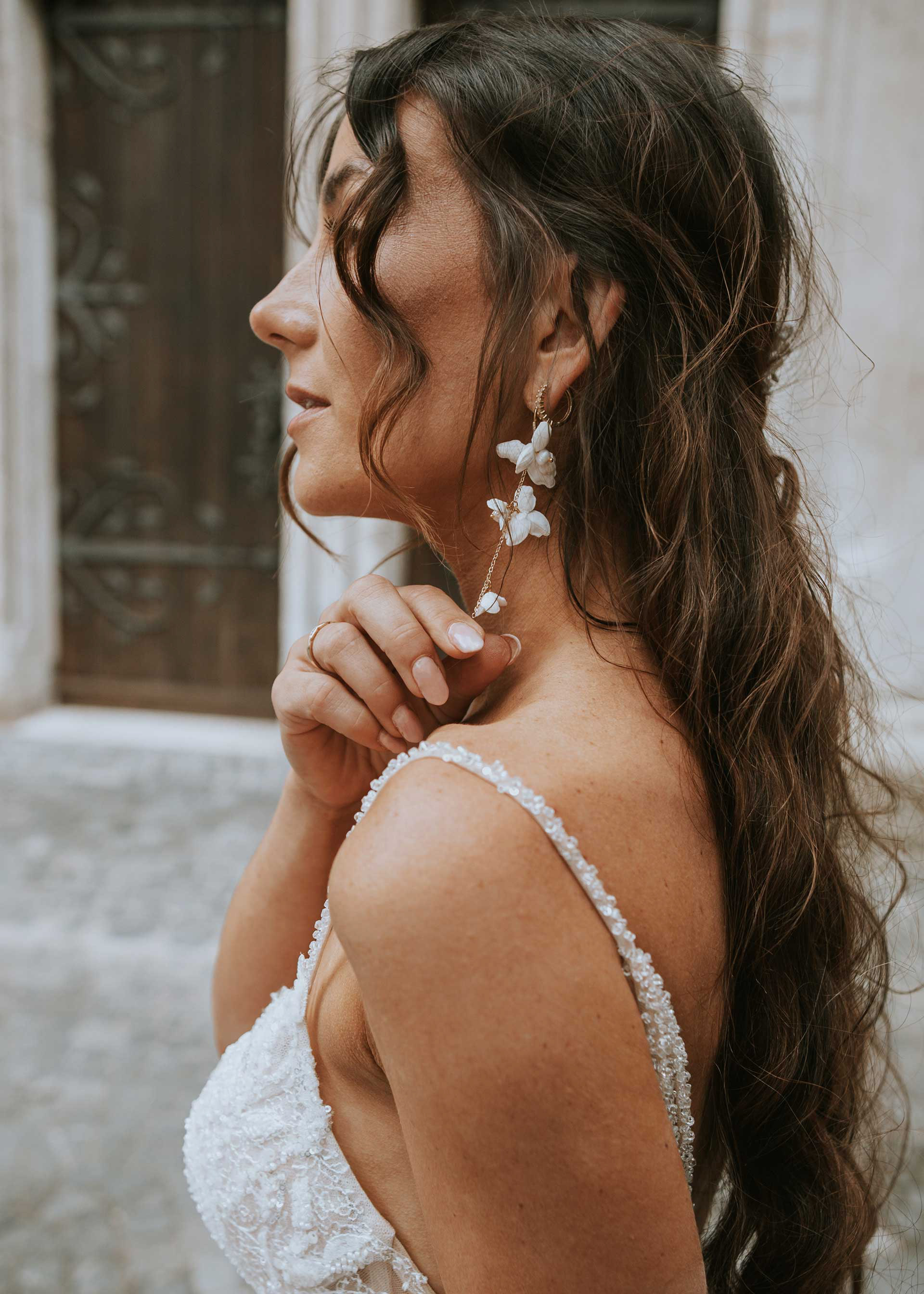 Seitenprofil einer Braut mit langem, lockigem Haar, die florale Ohrringe und ein besticktes Hochzeitskleid trägt. Ihre Hand mit einem Ehering ist an ihrem Hals zu sehen.