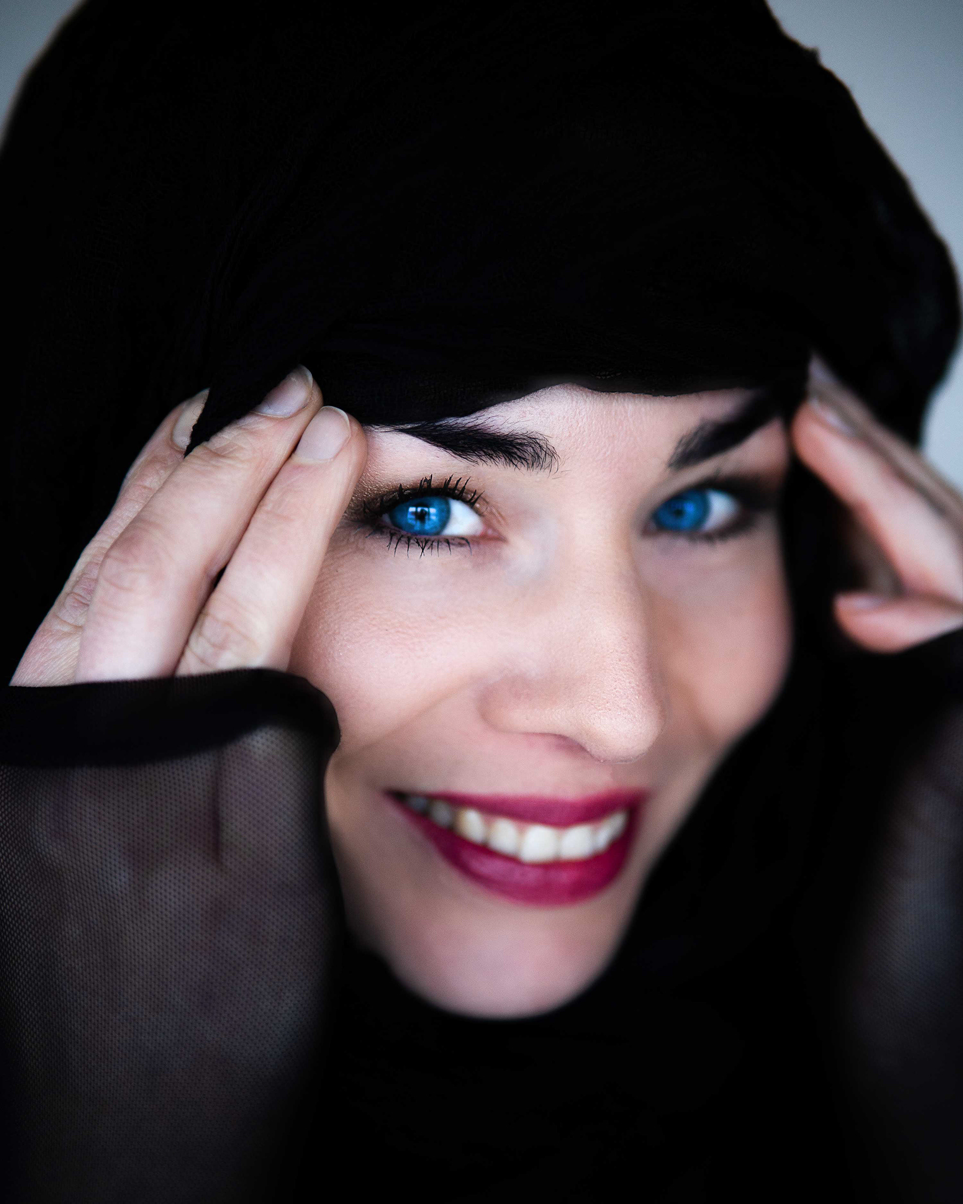 Detailaufnahme eines Gesichts mit intensivem Blick, blauen Augen und dunklem Lippenstift, umhüllt von schwarzem Schleier, von Medusa Fotografie Regensburg
