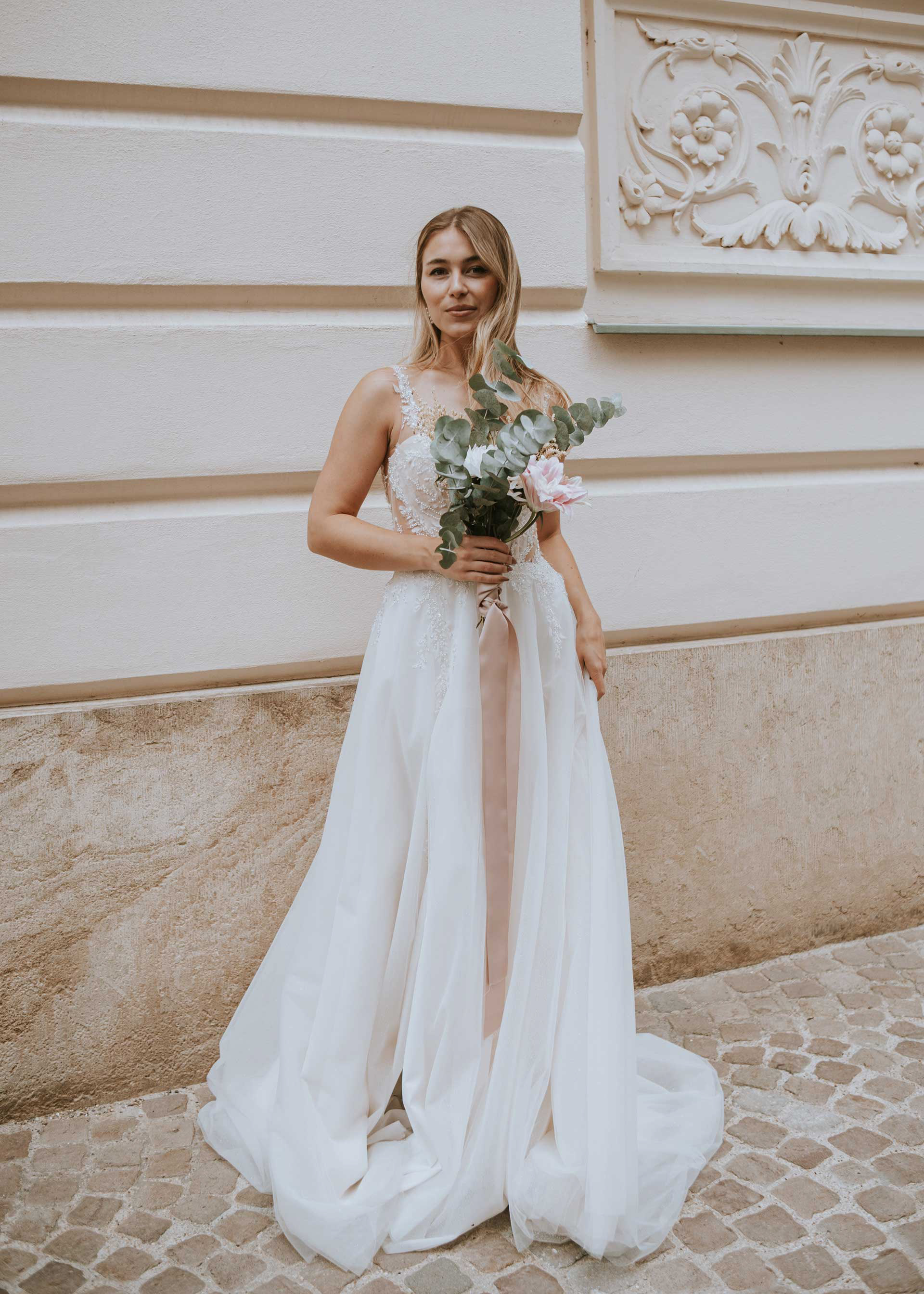 Ganzkörperaufnahme einer eleganten Braut vor einer historischen Regensburger Fassade mit Stuckrelief. Sie präsentiert ihr A-Linien-Brautkleid und den modernen Eukalyptus-Brautstrauß mit langem Satinband. Foto: Medusa Fotografie
