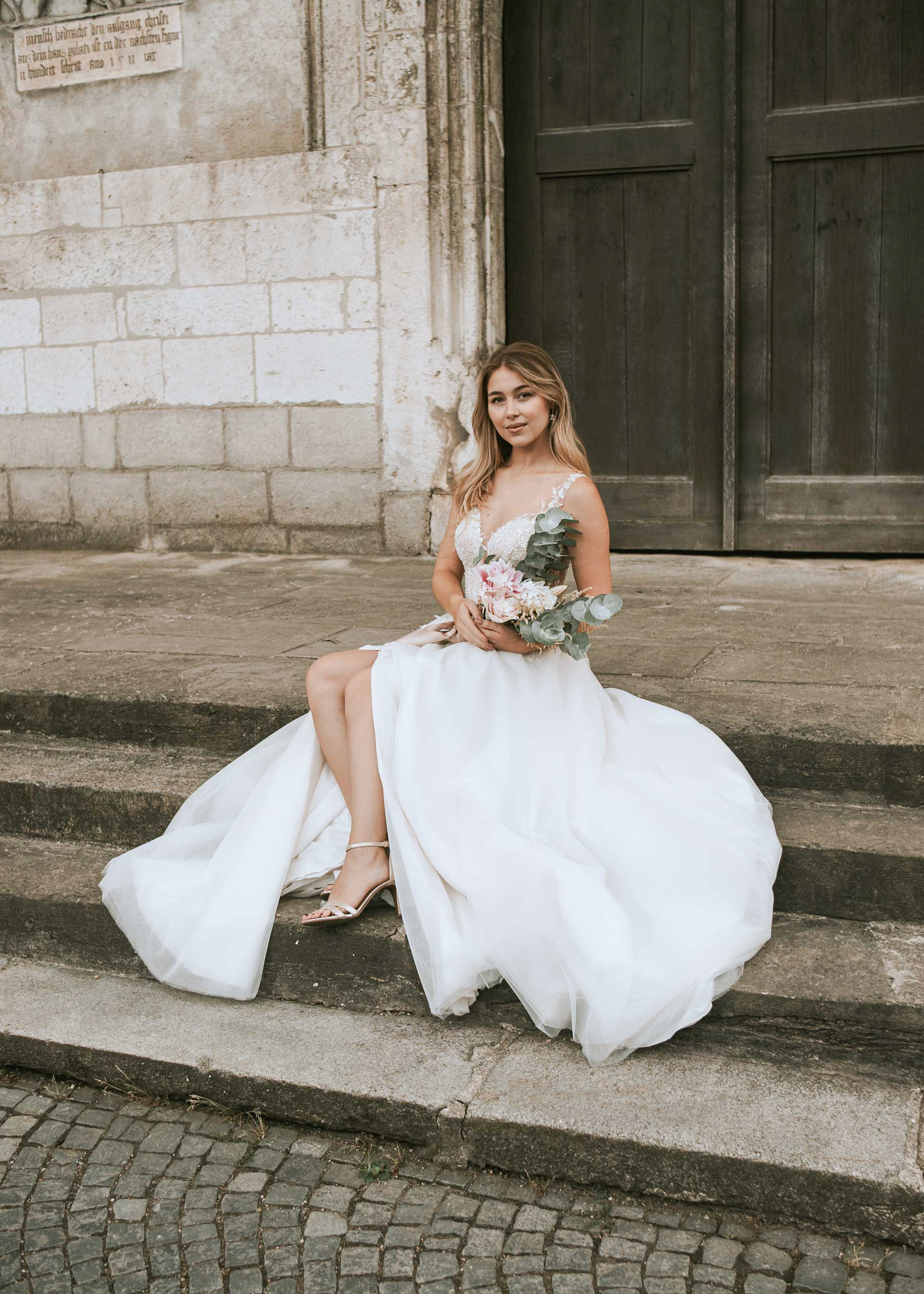 Atmosphärisches Foto von Medusa Fotografie: Die Braut sitzt elegant auf den Steinstufen vor der historischen St. Emmeram Basilika in Regensburg. Das Brautkleid mit Beinschlitz und der Eukalyptus-Strauß sind sichtbar