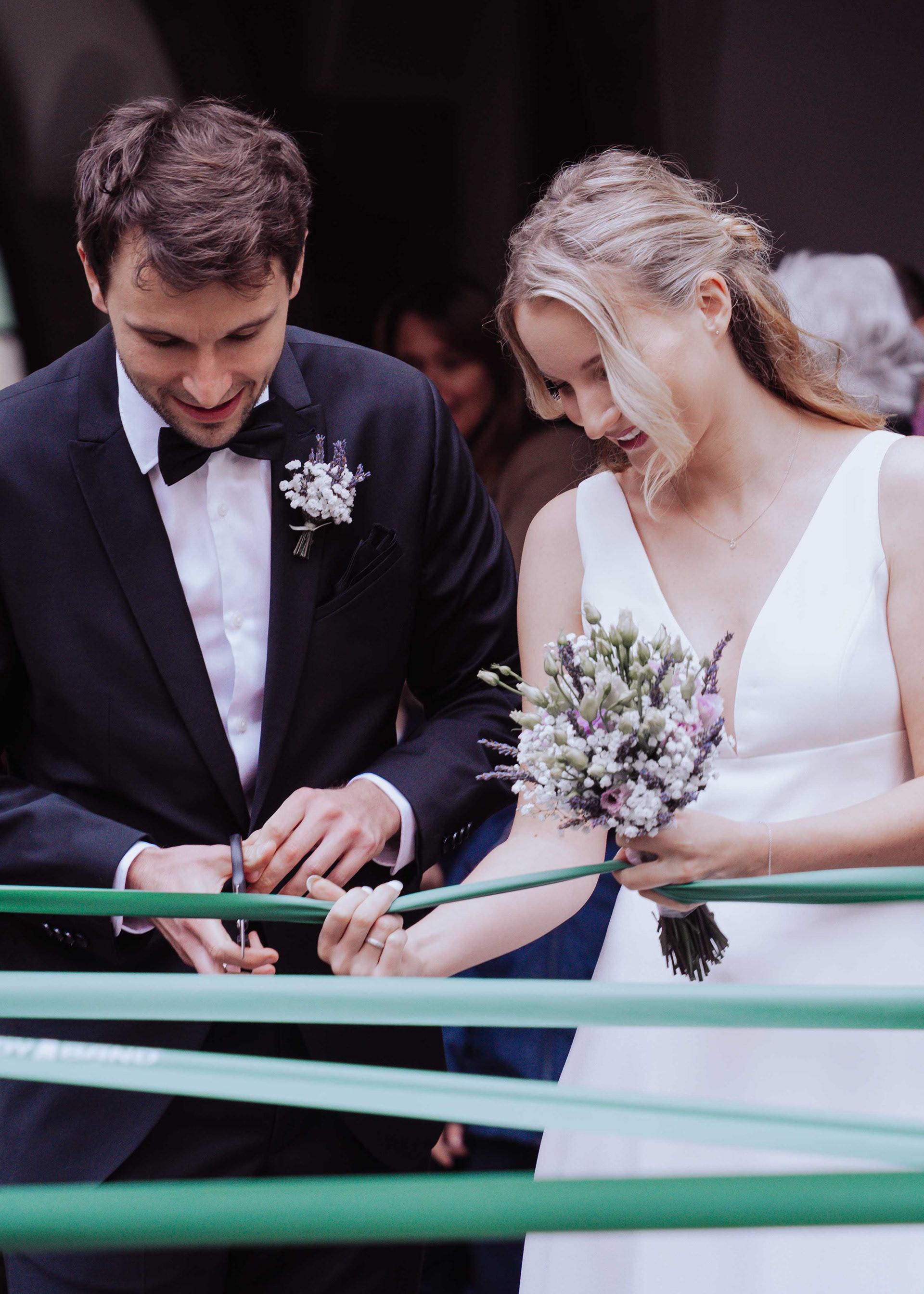 Nahaufnahme eines Brautpaares, das gemeinsam ein grünes Band durchschneidet. Der Bräutigam im schwarzen Anzug mit Fliege und der Lavendel-Boutonnière, die Braut im weißen V-Ausschnitt-Kleid mit ihrem Brautstrauß. Professionelle Hochzeitsfotografie Regensburg von Medusa Fotografie.