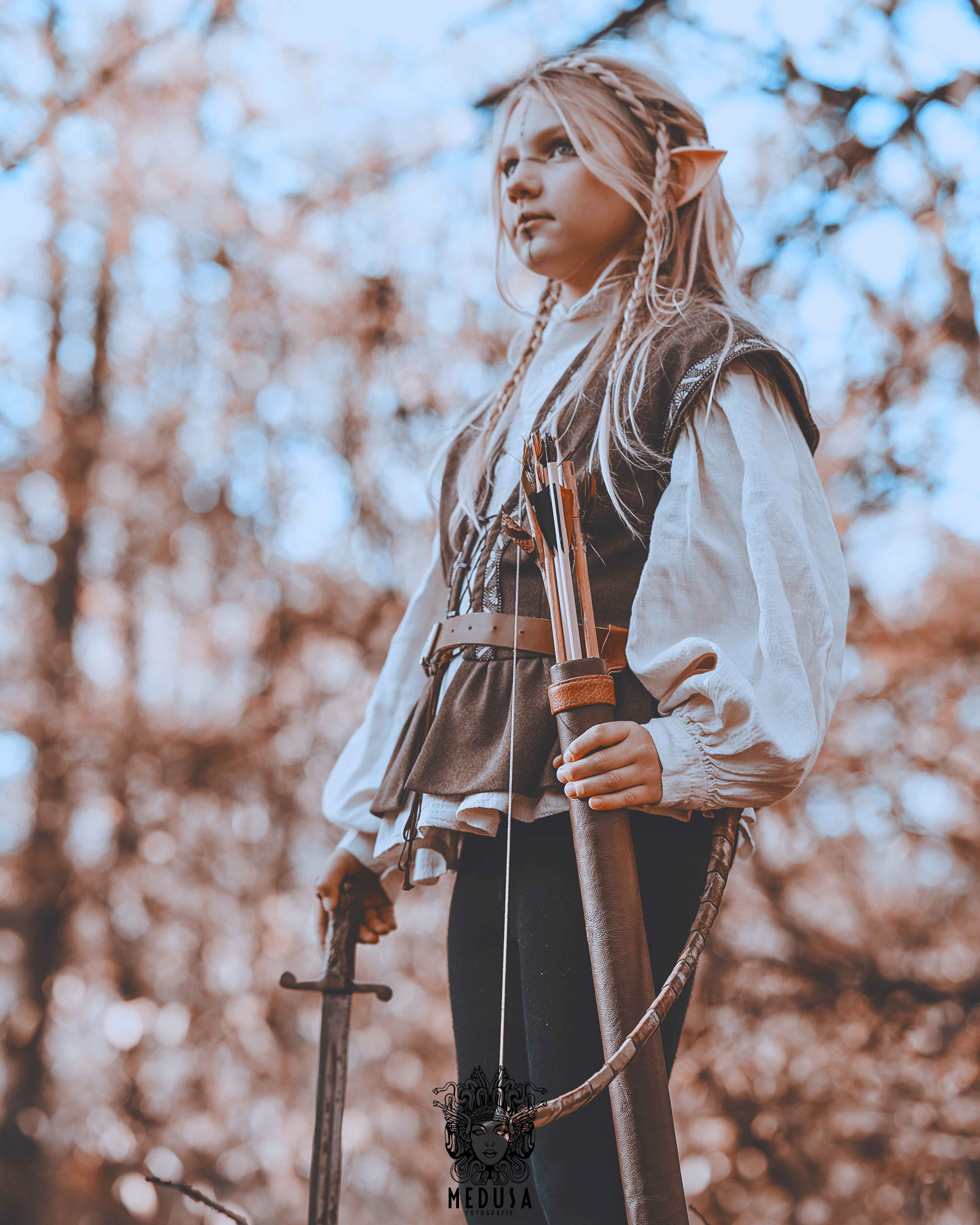 Halbporträt einer bewaffneten Elfen Kriegerin, die einen Bogen hält, fotografiert im herbstlichen Wald. Professionelle Cosplay Fotografie von Medusa Fotografie mit stimmungsvollem Bokeh-Effekt und cineastischem Film-Look