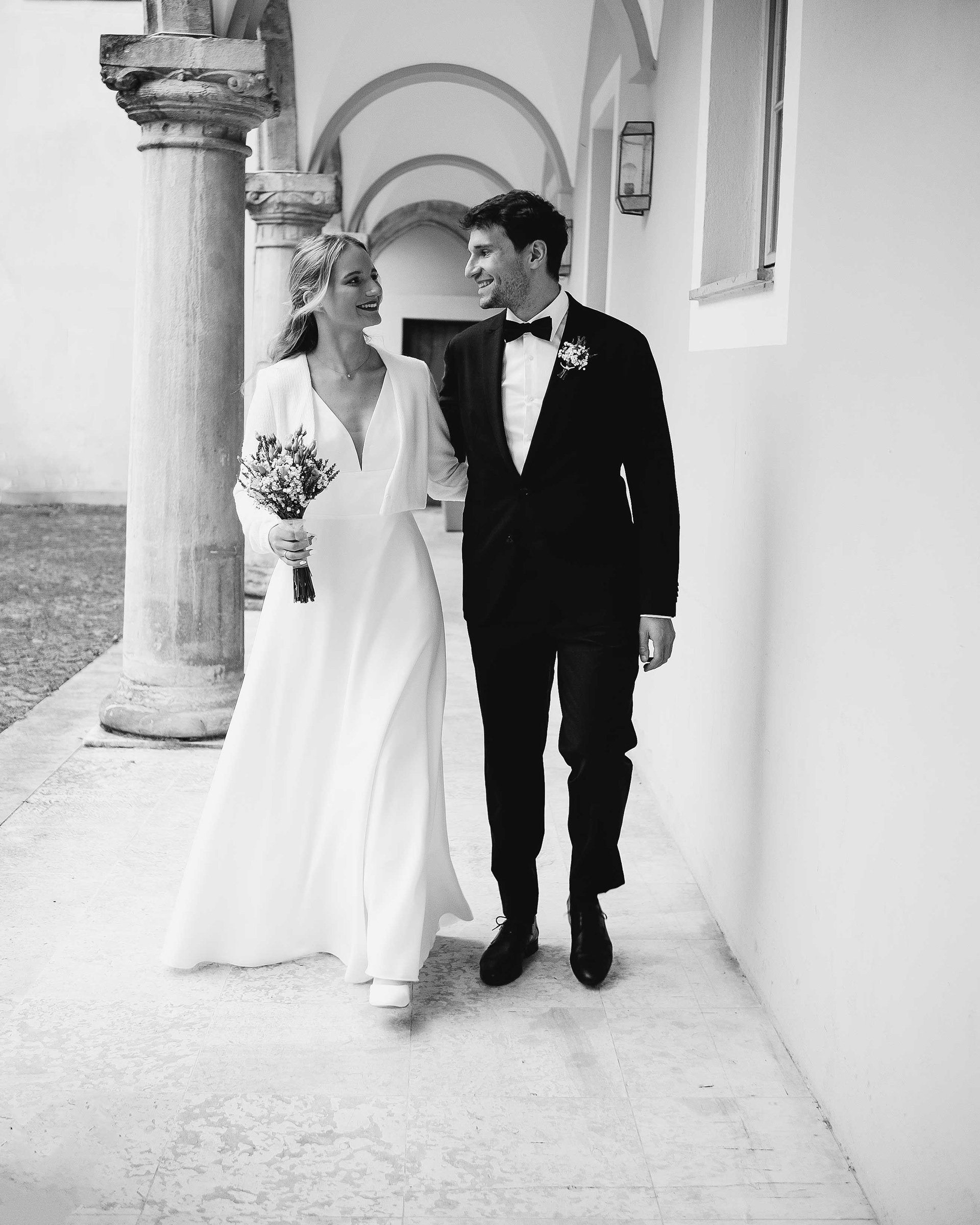 Schwarz-Weiß-Fotografie eines frisch vermählten Brautpaares, das lächelnd durch einen historischen Bogengang in Regensburg spaziert. Die Braut trägt ein schlichtes weißes Hochzeitskleid mit Jäckchen und hält einen Brautstrauß, der Bräutigam einen klassischen dunklen Anzug mit Fliege.