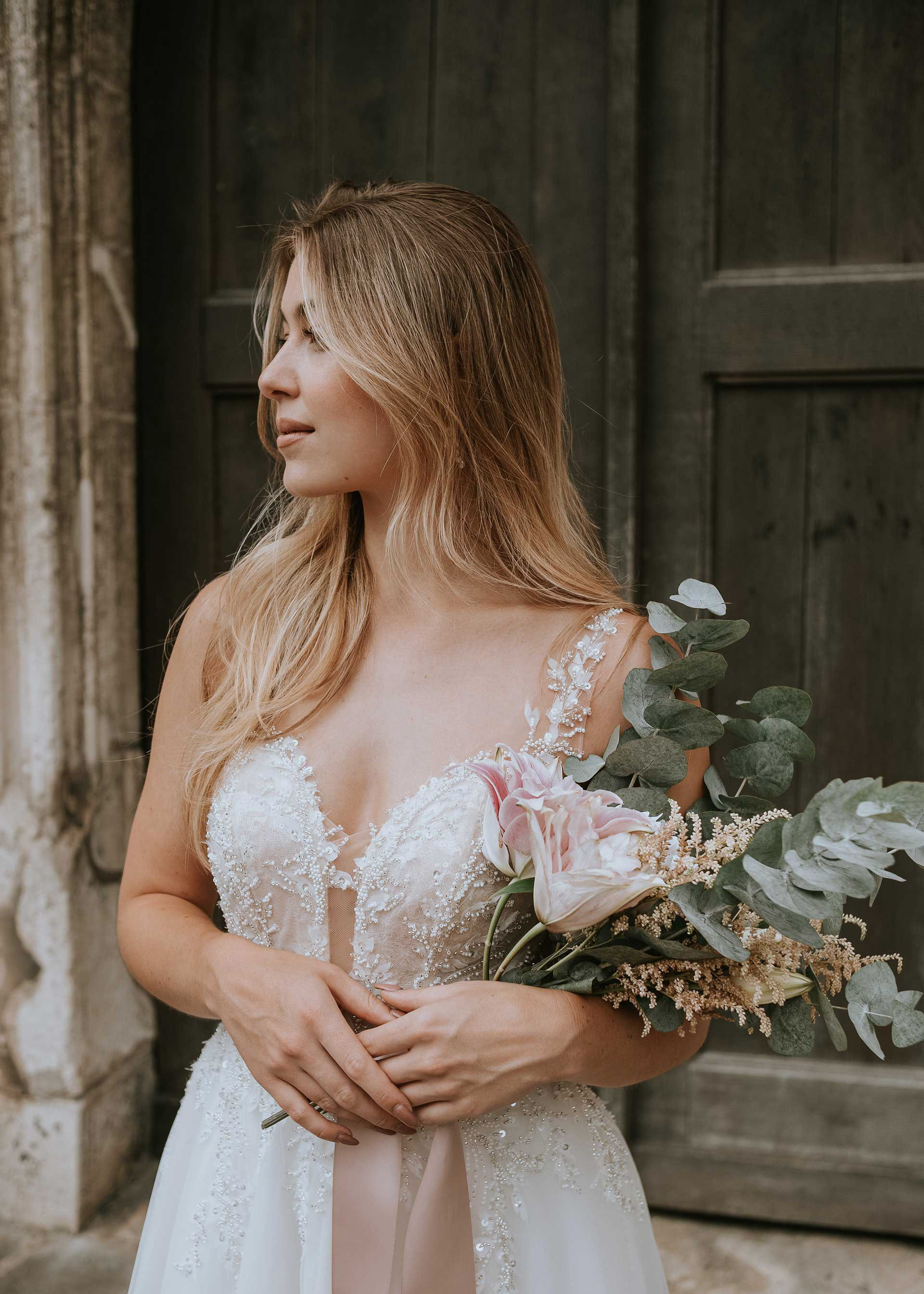 Elegantes Brautshooting-Foto vor dem historischen Portal der Basilika St. Emmeram in Regensburg. Die Braut steht im Spitzenkleid vor dem dunklen Holztor und blickt zur Seite. Medusa Fotografie.