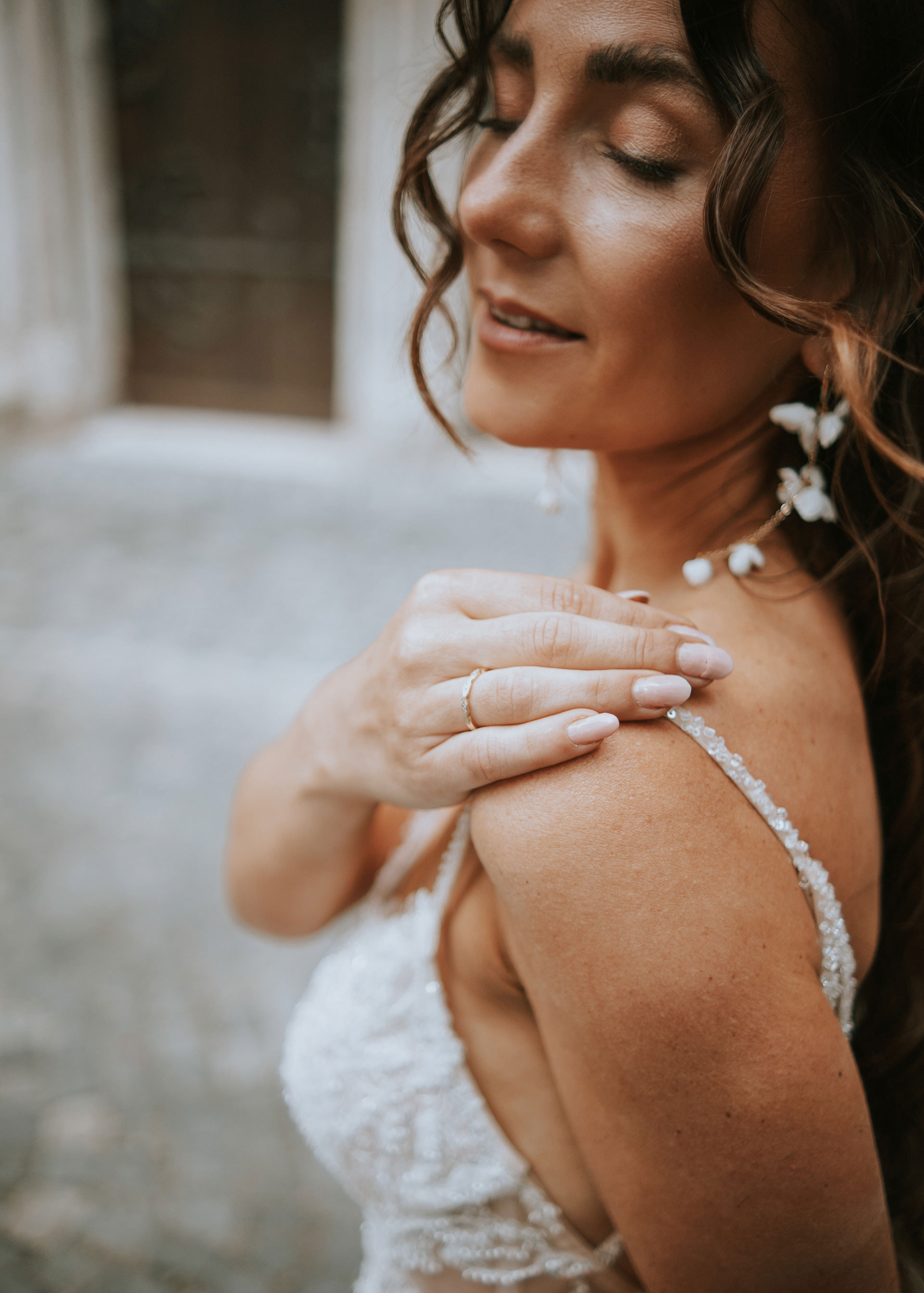 Nahaufnahme einer Braut in einem bestickten Hochzeitskleid, die eine Hand mit einem Ehering auf ihre Schulter legt. Die Braut trägt außerdem florale Ohrringe und hat ihr Haar hochgesteckt.