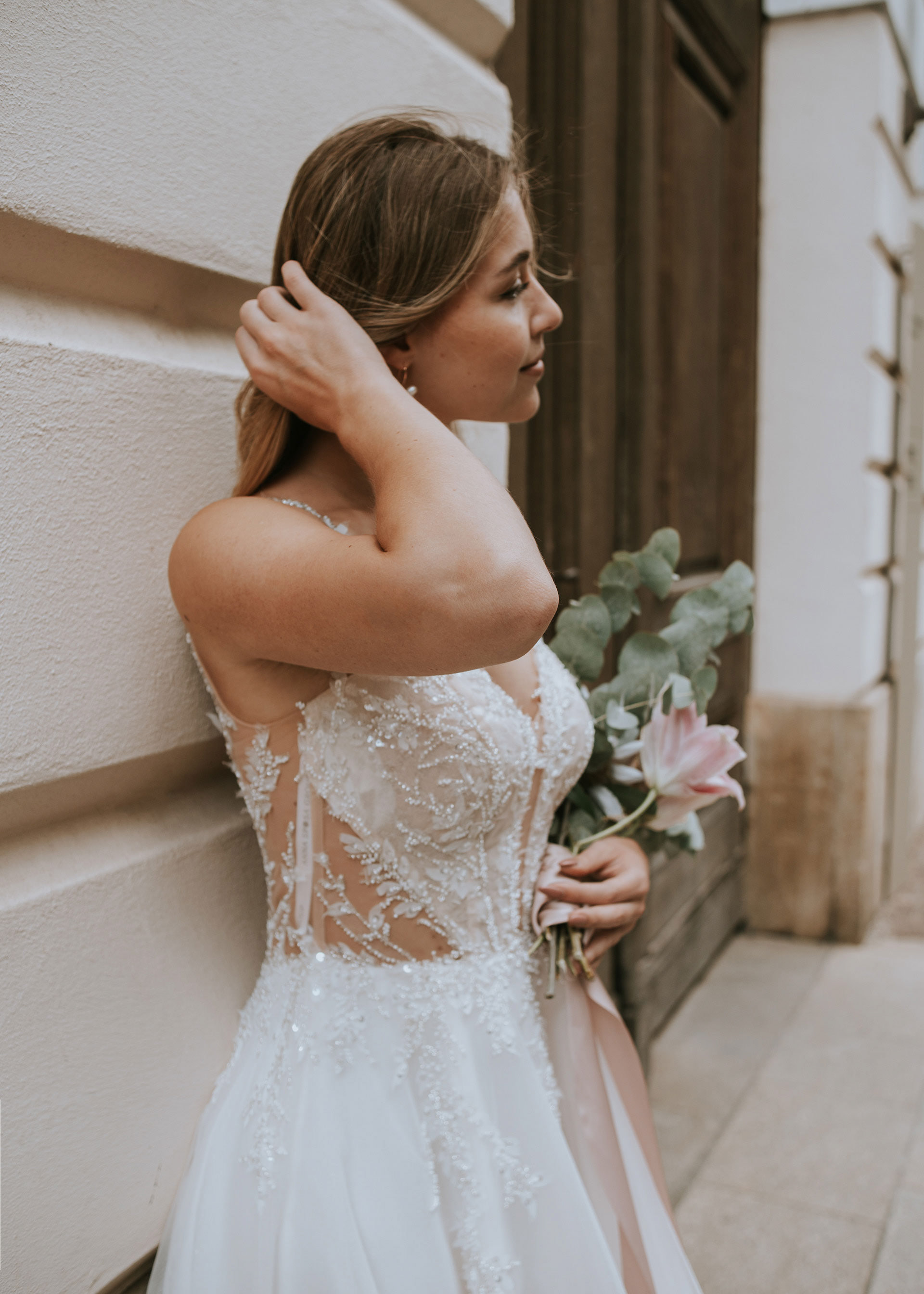 Seitenprofil-Porträt einer Braut beim Shooting in Regensburg mit Medusa Fotografie. Die Braut blickt an der stuckverzierten Fassade vorbei, hält ihren Hochzeitsstrauß und richtet sich das blonde Haar.