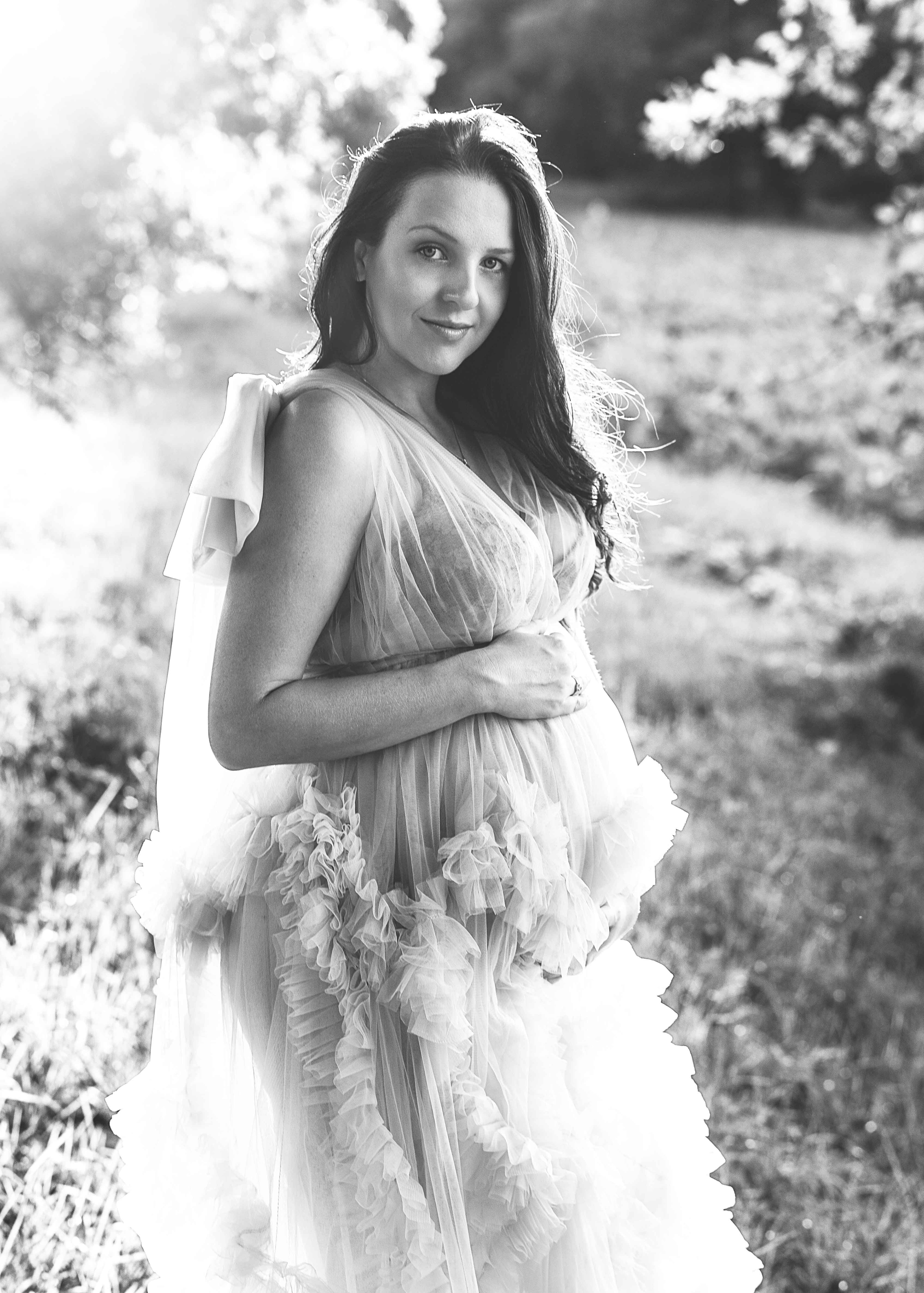 Ein verträumtes Schwarz-Weiß-Foto einer werdenden Mutter in einem opulenten, hellen Tüllkleid, das durch das sanfte Gegenlicht der Sonne in der Natur zum Leuchten gebracht wird.
