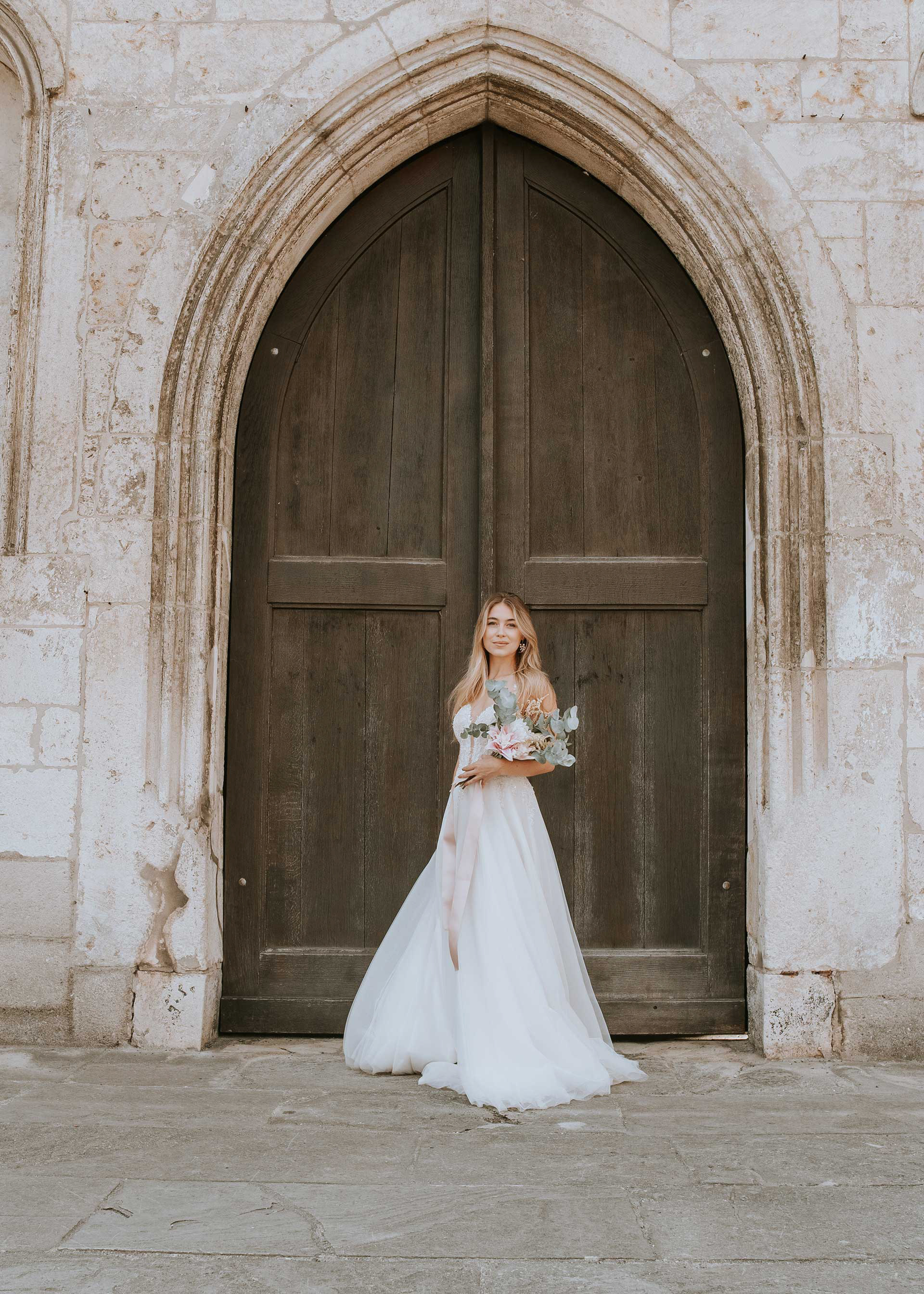 Weitwinkelaufnahme der Braut im imposanten, gotischen Spitzbogen-Portal der St. Emmeram Kirche Regensburg. Die Architektur rahmt die Braut ein. Foto: Medusa Fotografie
