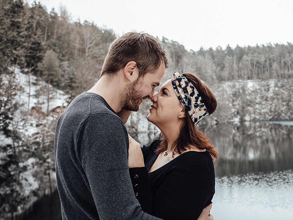 Authentischer Moment beim Outdoor-Fotoshooting, bei dem der Mann die Frau von hinten umarmt und beide glücklich am Seeufer lachen