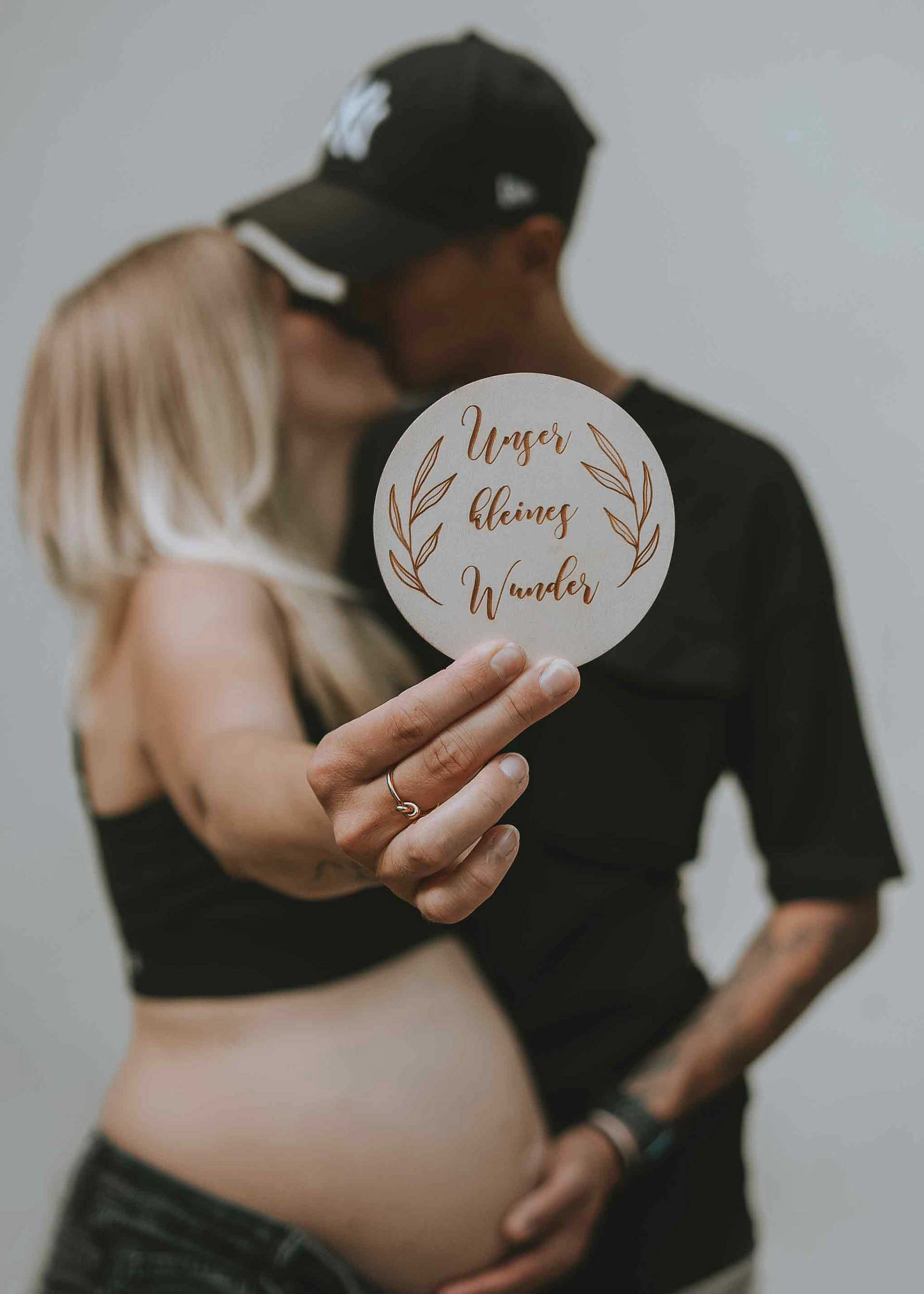 Detailaufnahme einer hölzernen Plakette mit der Aufschrift Unser kleines Wunder vor einem küssenden Paar mit Babybauch