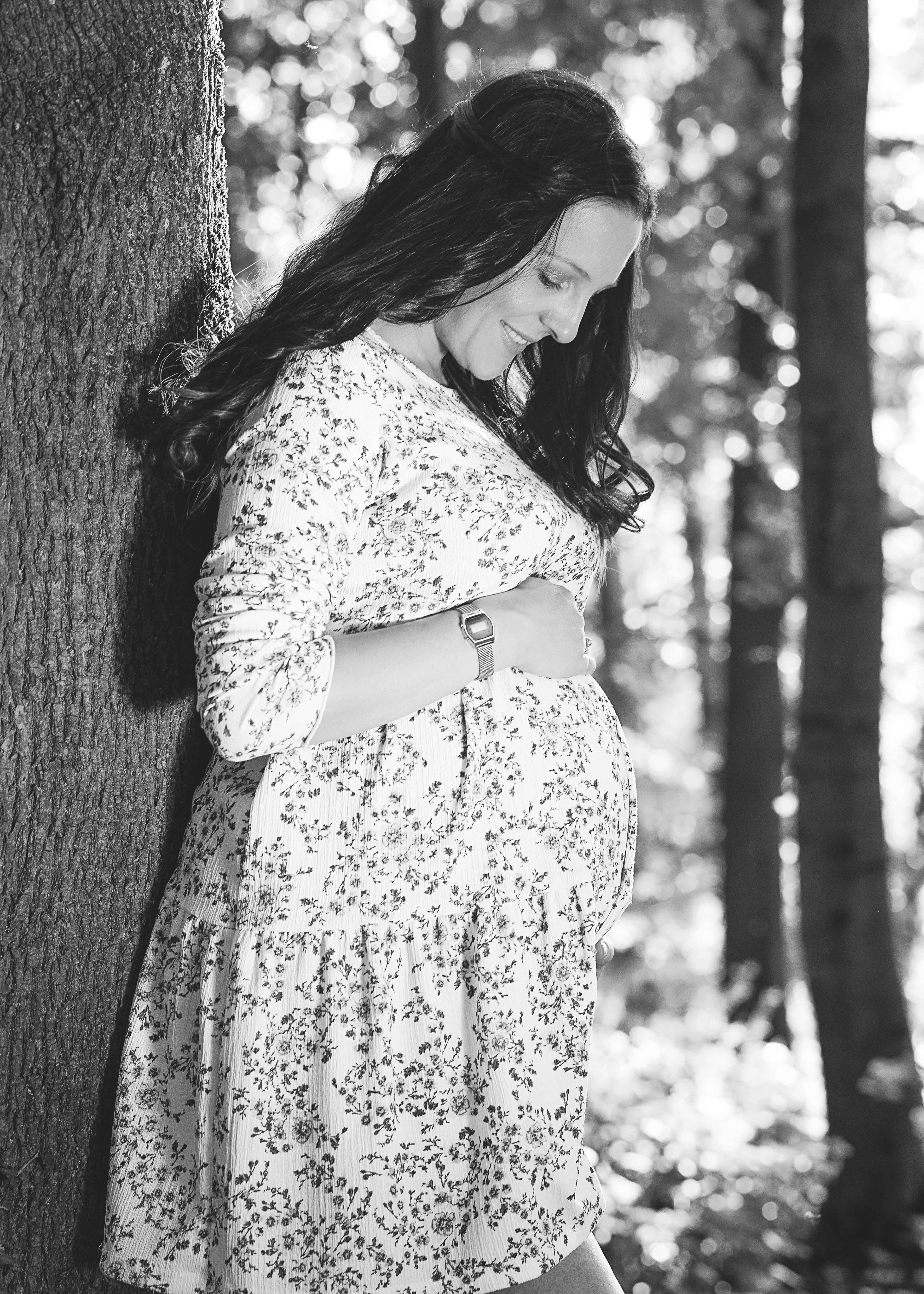 Ein emotionales Schwarz-Weiß-Porträt einer schwangeren Frau im floralen Kleid, die sich lächelnd an einen Baumstamm lehnt und behutsam ihren Babybauch hält.