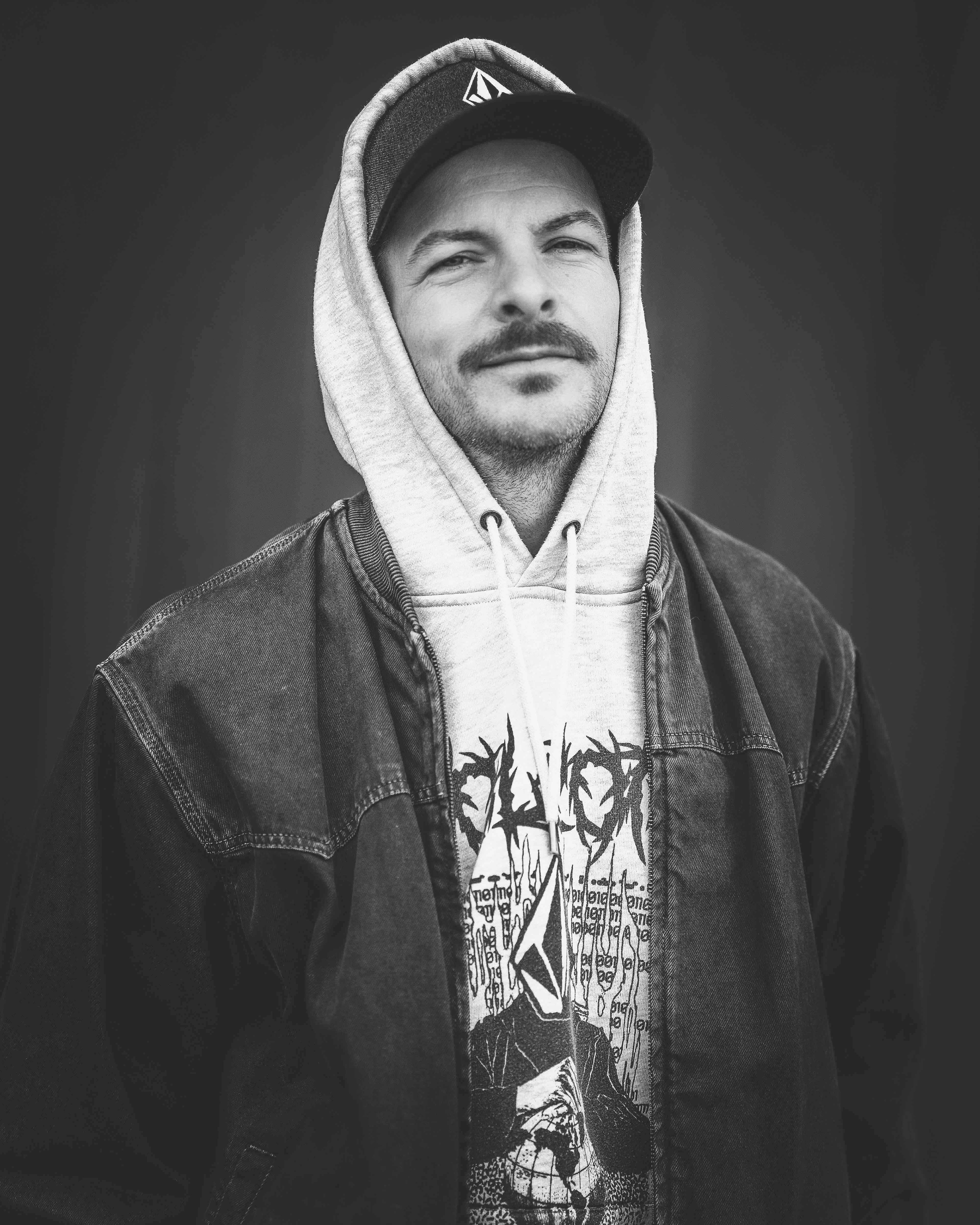 Schwarz-Weiß-Porträt eines Mannes mit Schnurrbart, Kapuzenpullover und Kappe, aufgenommen im Hafengebiet Regensburg von Medusa Fotografie