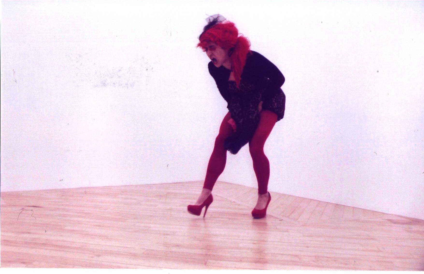 This is from a performance in Boston. It is also a grainy image and was taken with a film camera. I am wearing the same black lace dress except I am wearing a red wig and red tights with red stiletto shoes. The background is white and the floor is a beautiful wood pattern. I am screaming and my hands are bunching my dress in my crotch area.