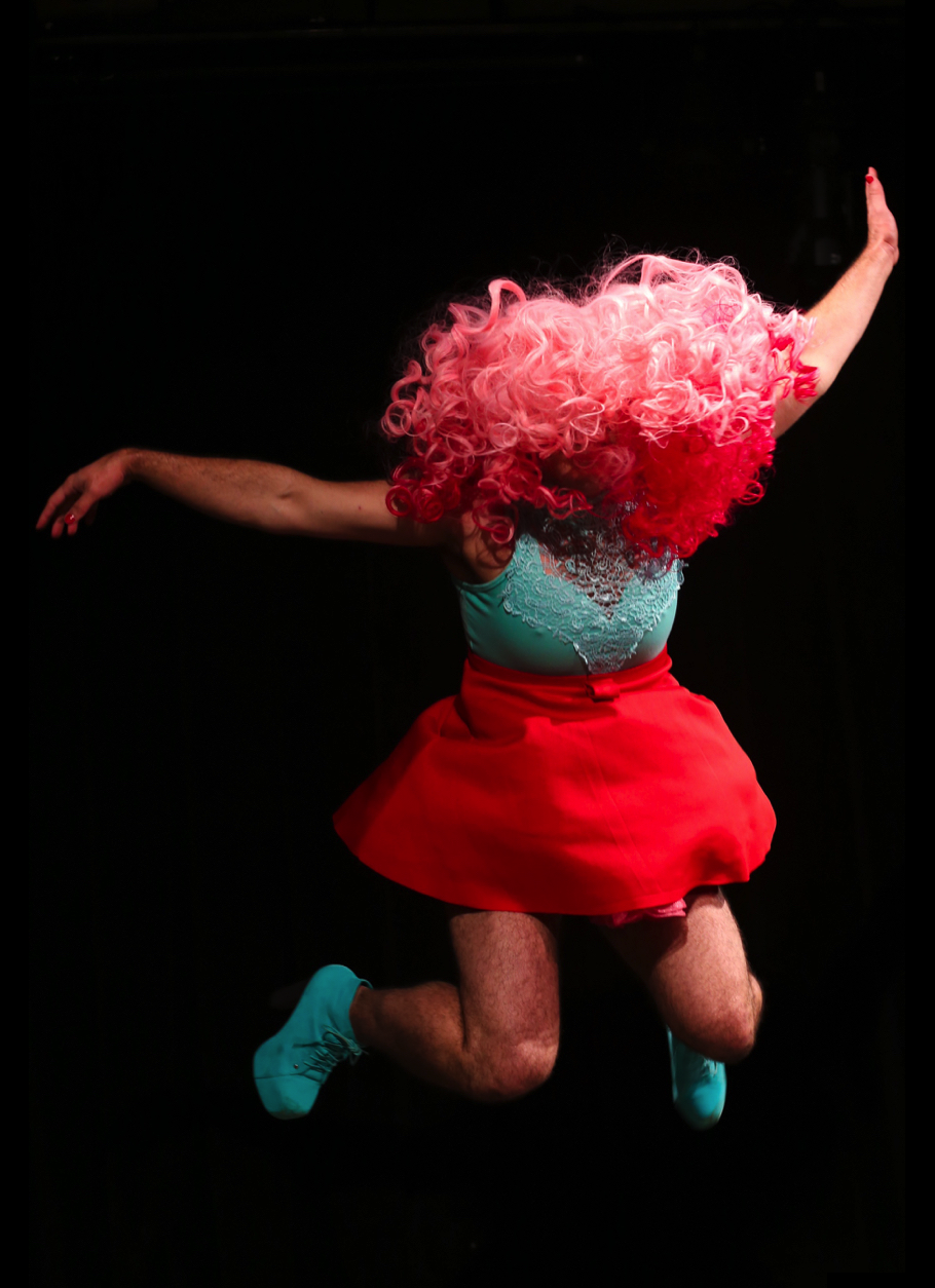 The following eight images are documentation from the performance I'm Fine in Brazil. I am performing against a black backdrop. I am wearing a bright red mini skirt, a teal halter top with a sweetheart neckline that has a lace applique over the front. I have on a long pink wig that is curly and fades from light pink to dark pink. I am wearing teal booties that are six inches high. I am mid jump and my entire body is off the ground. My legs are hairy and so are my armpits.