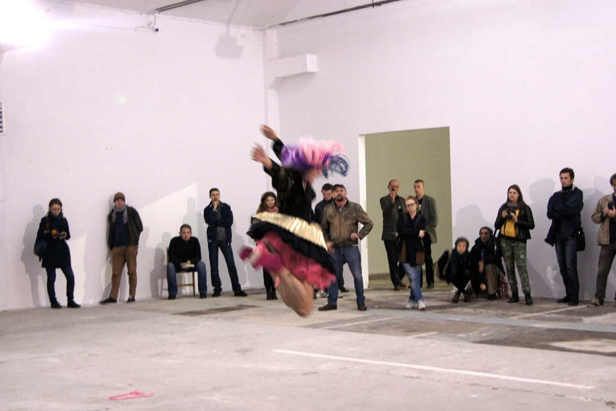 The next six images are from a performance I did in Lublin Poland for Performance Platform in 2013. I am wearing a long wig that fades from light pink to purple to blue. I am wearing a black halter top and a crop top jacket that has a subtle pattern of gold glitter flowers. I am wearing a pleated gold extra mini skirt with a pink tutu underneath. My shoes are pink platform booties with a six inch heel. You can see the hair on my legs. The crowd is big and the space is huge. It is a bright space with white walls and a concrete floor. Most of the images are pretty far away from the camera's lens.