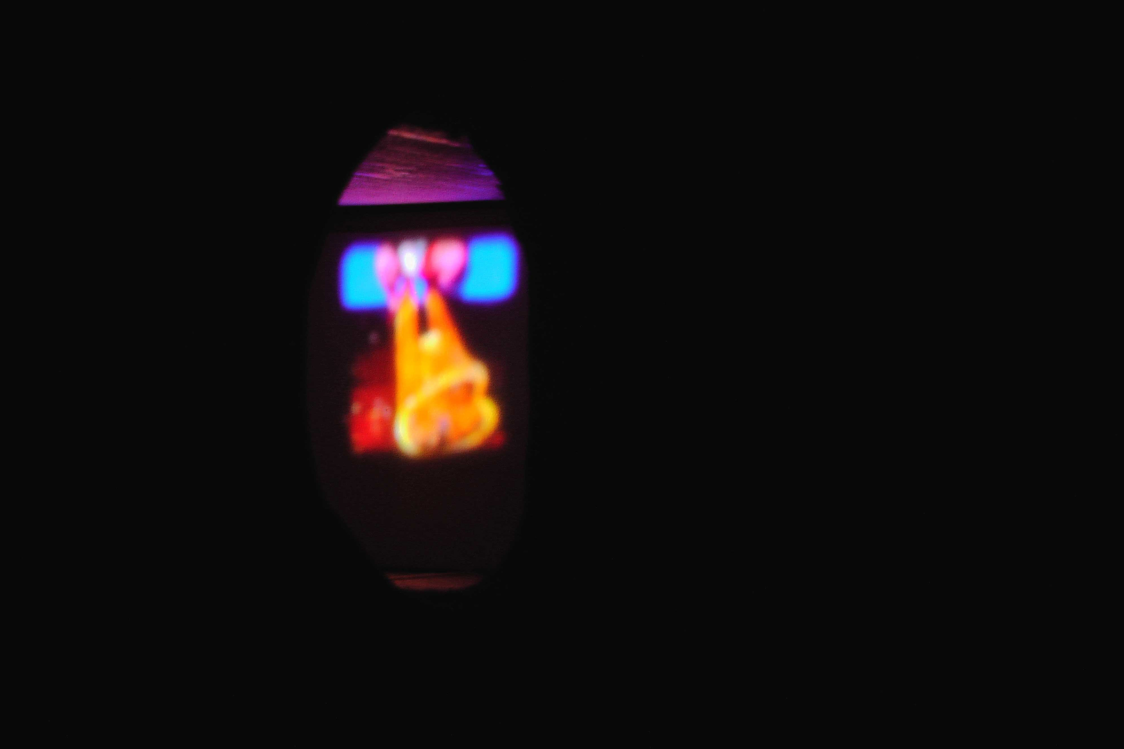 A screenshot of the artist as Pinocchio. They are in a Pinocchio costume with puppet strings and white gloves. The majority of the image in black and in the center of the image is a small peep hole where the image appears.The artist is sitting on a large painted yellow log; only the bottom half of their body is in the image.
