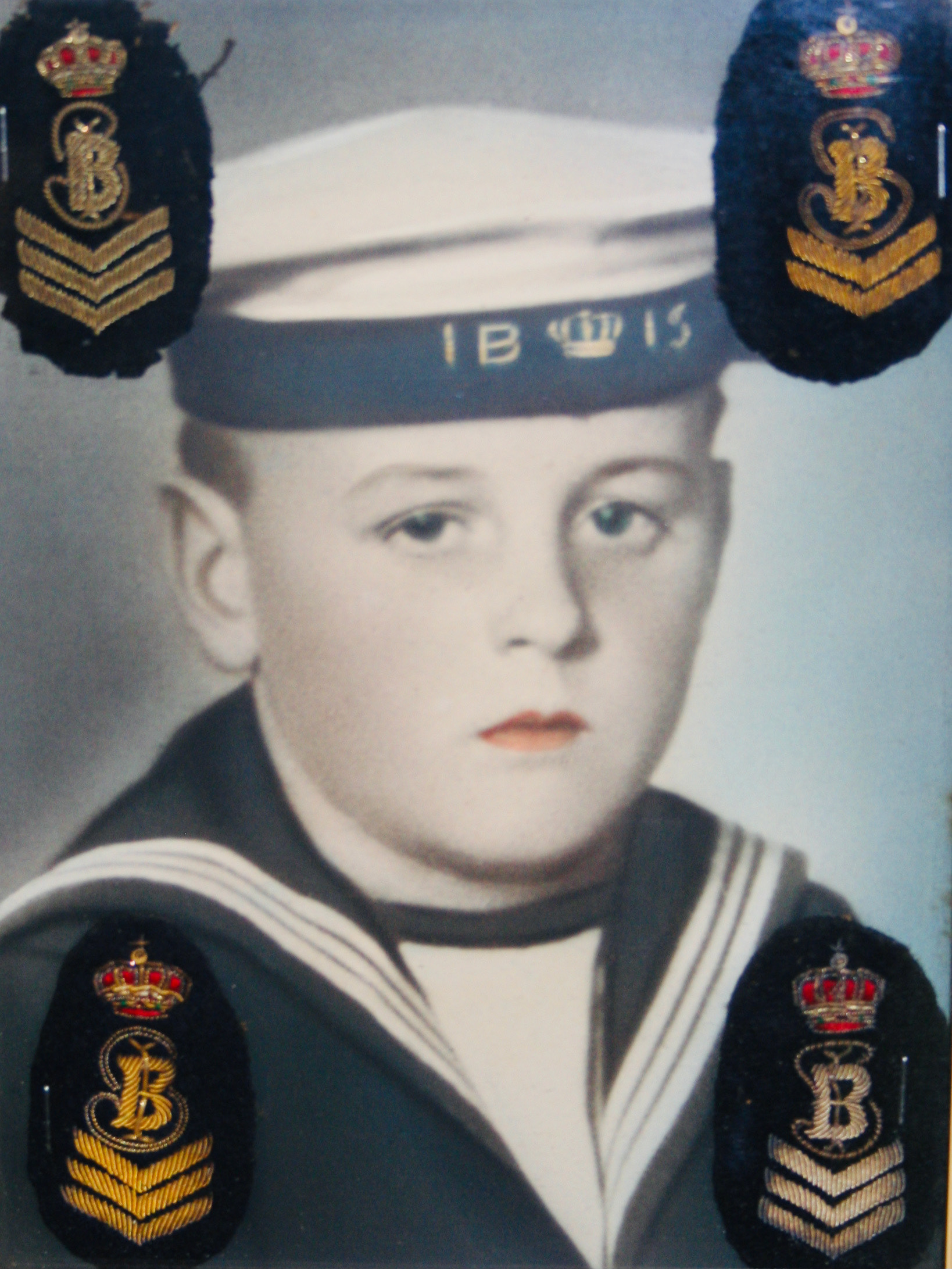 a hand-coloured black and white identity portrait of a boy in a sailor suit with blue eyes and orange lips. On his sailor hat it says IBIS and the hat also has a crown, four emblems are sewn in the corners of the photograph