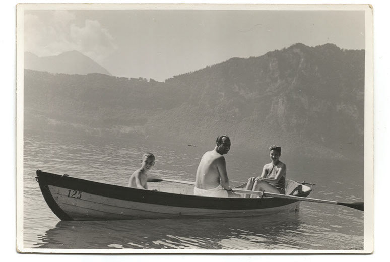 A black and white photograph from the 1930's of a rowing boat on a Swiss Lake with mountains in the background. In the boat, a young boy with his parents in swim clothing.