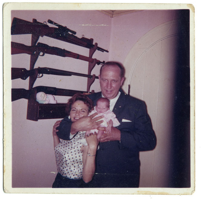 A vintage square colour photograph from the 1950s or early 1960s. A man in a black suit with white shirt shows a newborn baby in a corner of a room with four rifles against the white walls in the background: his wife is dressed in a white shun top with black polka dots and the newborn baby is dressed in woolly light pink cardigan. A figurine of a baby lies sleeping against the trigger of a shotgun. The tiny baby is a bonsai version of his (grand)father, with matching hairlines. The man holds his smiling wife in a strangling clasp.