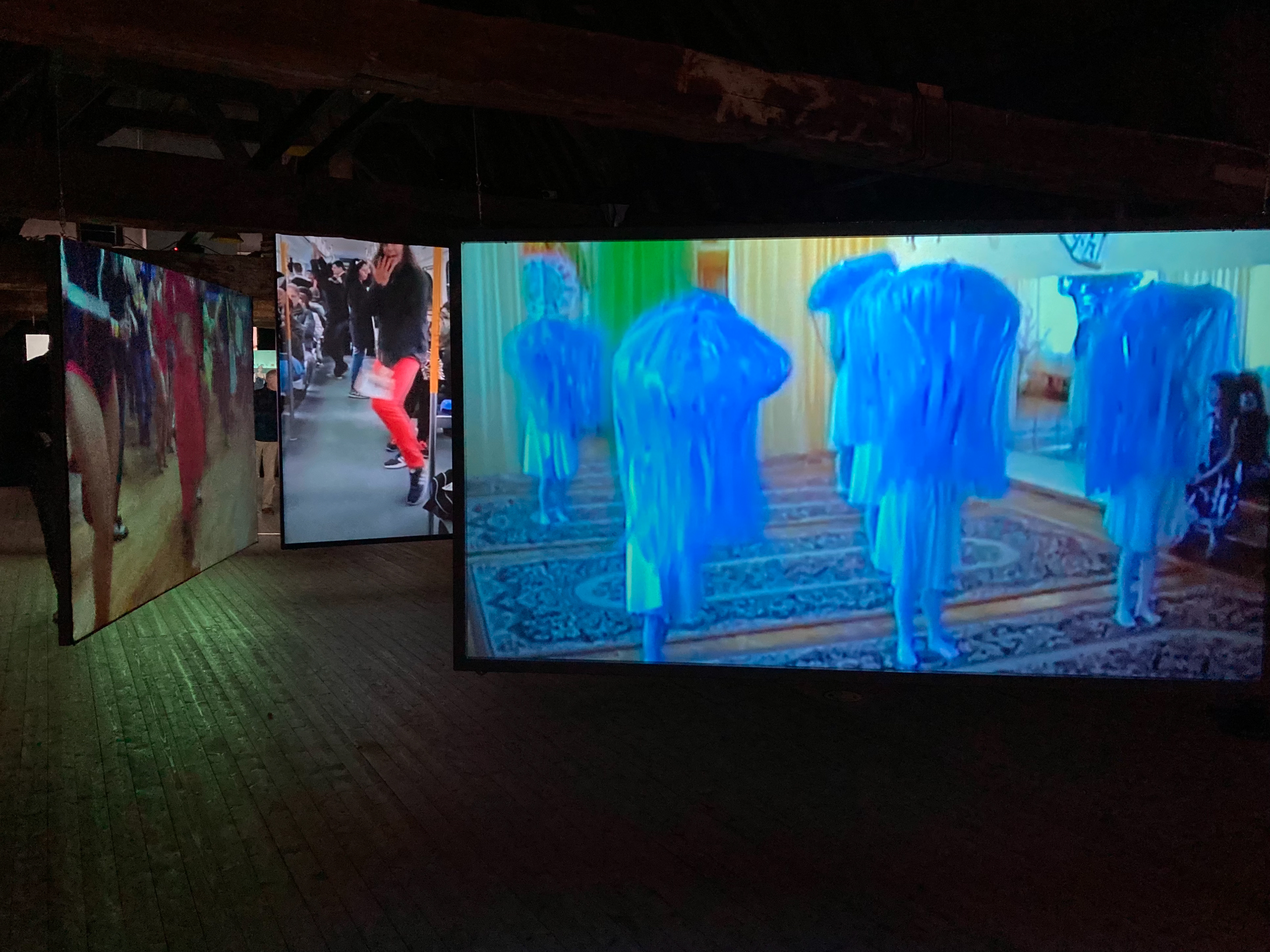A colour photograph of a darkened exhibition space with three screens,, each showing a different video of dancing people at various locations. the largest screen shows six dancing people wearing blue costumes which makes them look like sea anemones