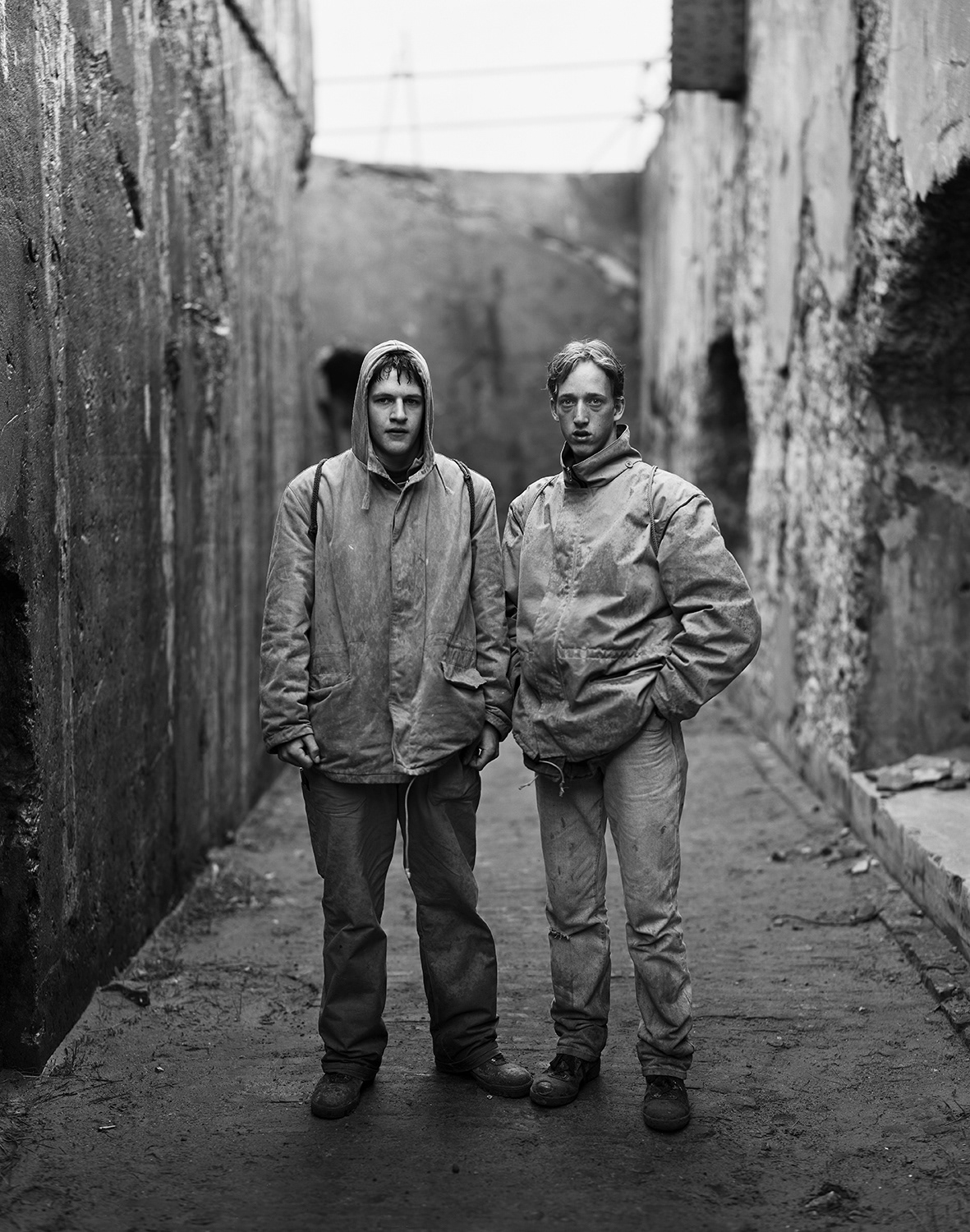 a black and white photograph of two young men in the rain wearing anoraks, jeans and working boots. these stand an a narrow passway with old dilapidated walls