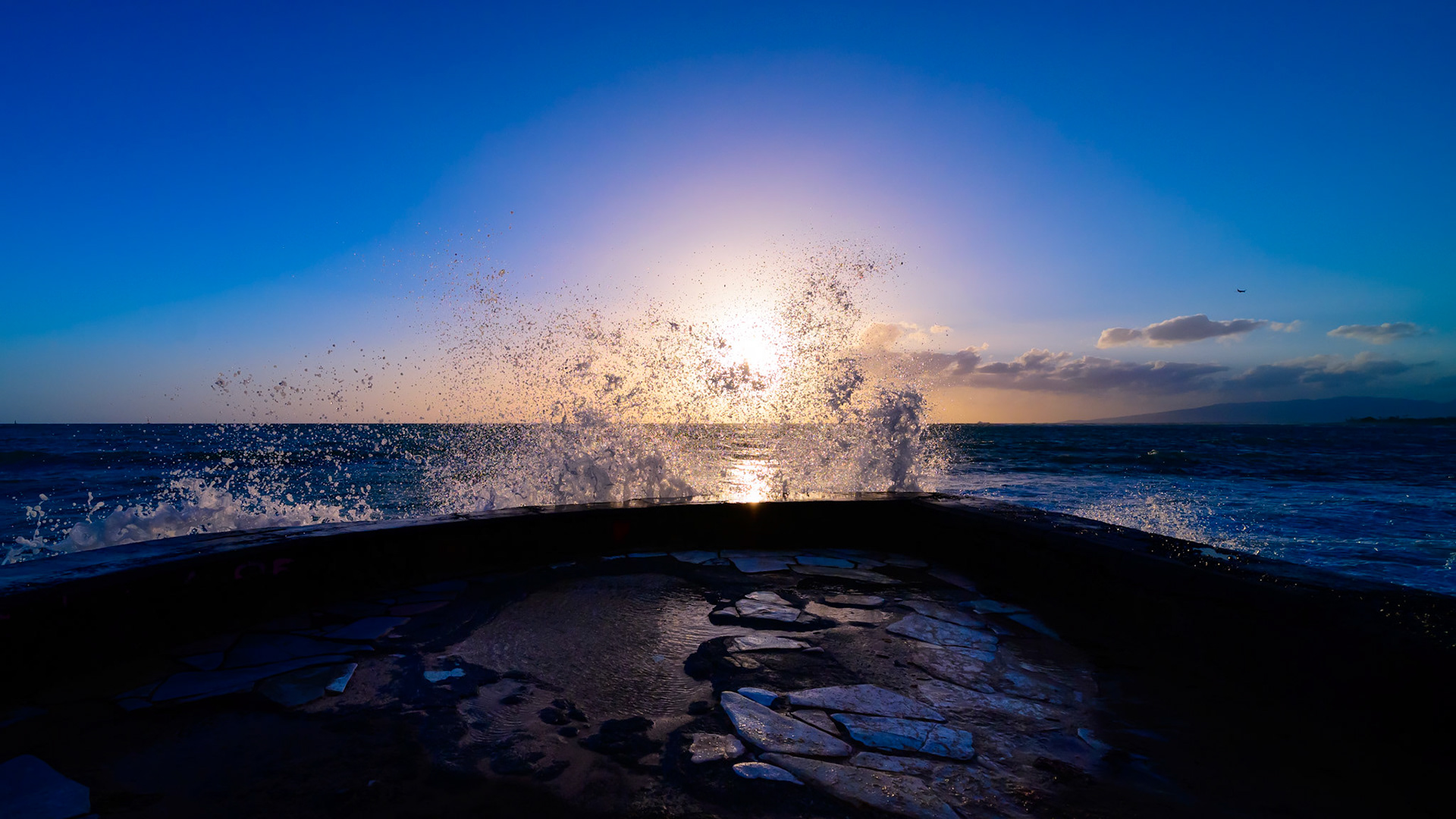 Wave splashes against the Waikiki Pier and the sunset light illuminates the water.