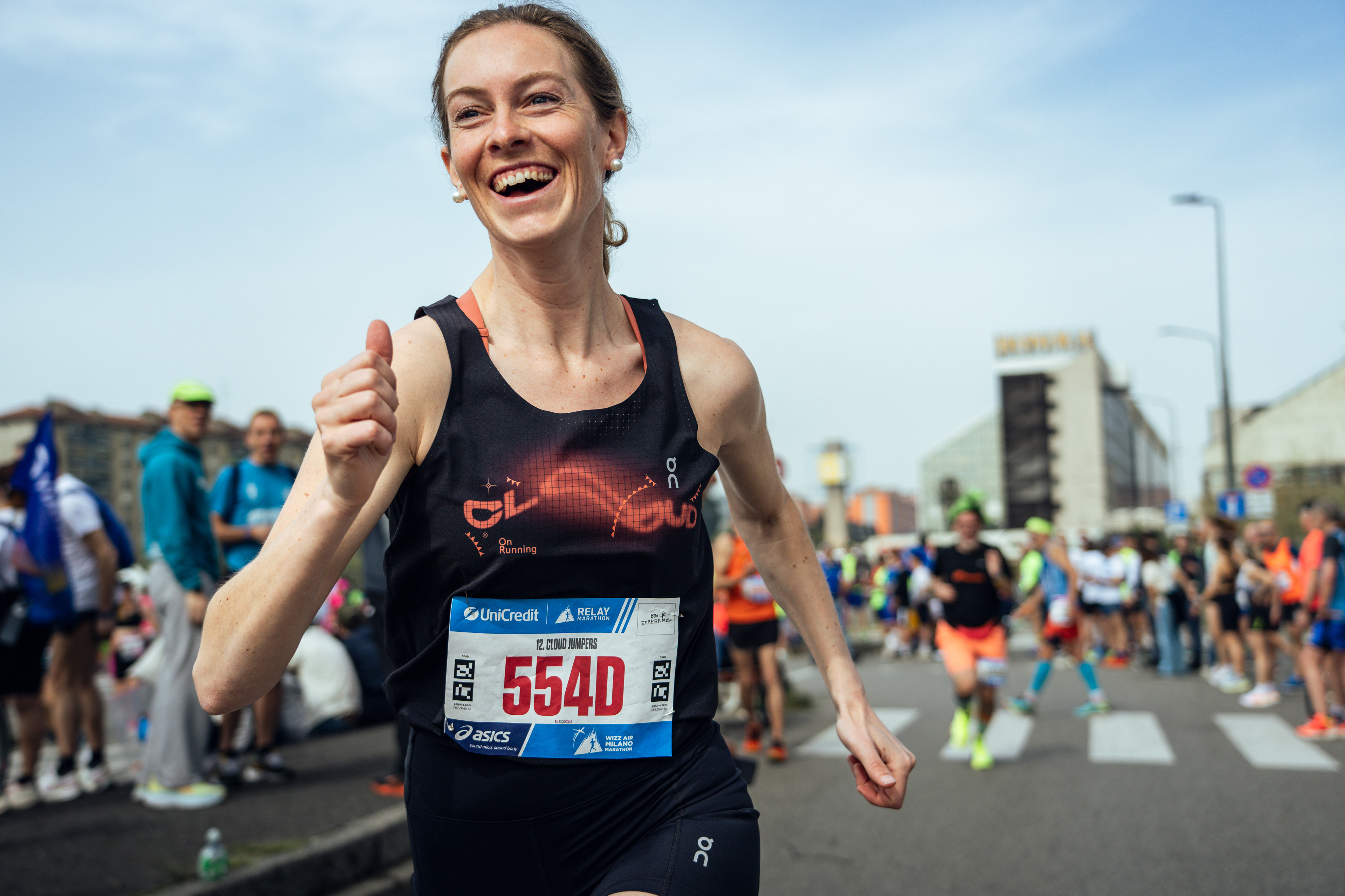 A smiling runner runs through a crowd lined street during the Milano Marathon. The sky is blue and there are a few buildings beyond the spectators.