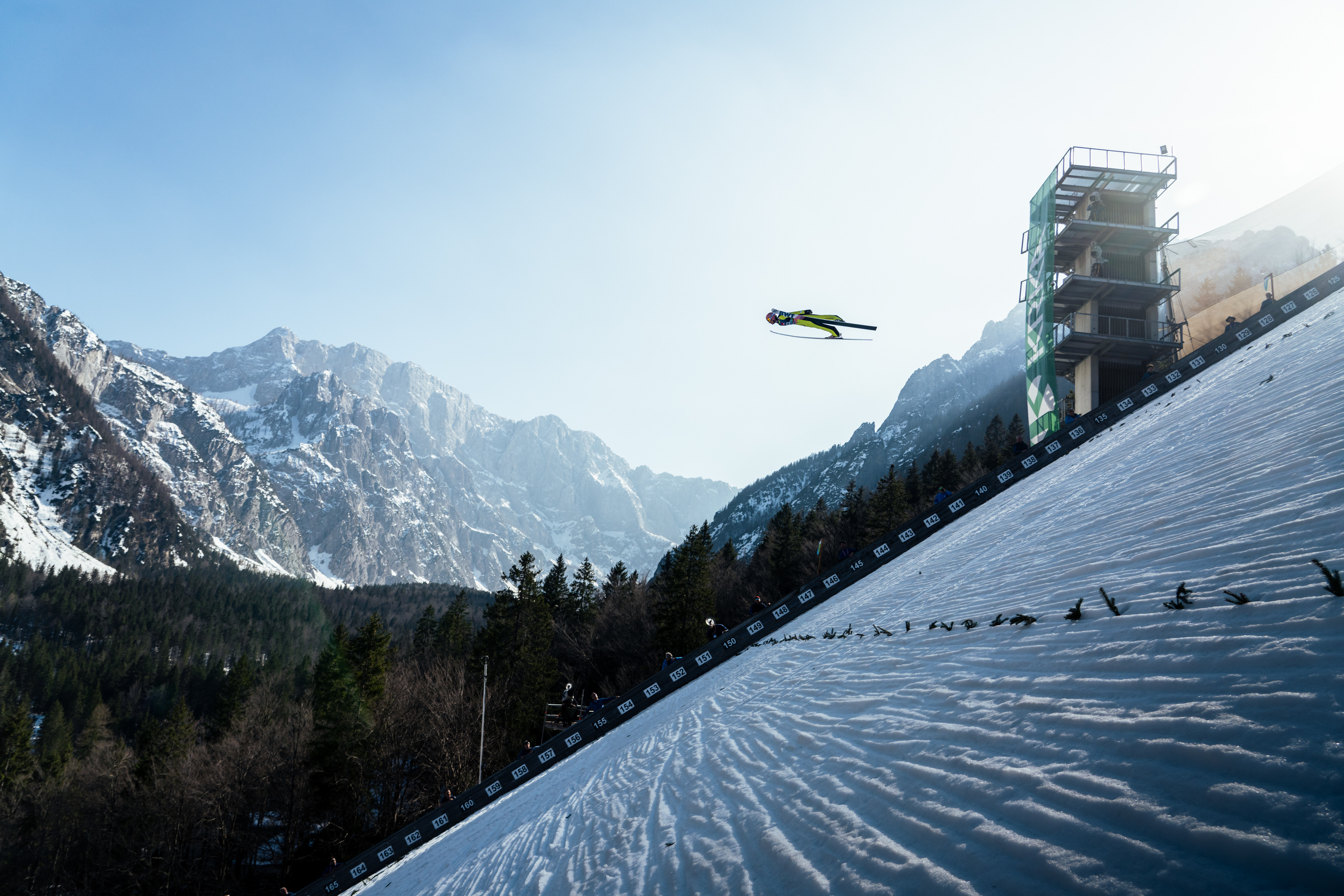 David Kobacki of Poland Flies very high above the ground in Slovenia. The lines of the snow grooming lead to the Ski Flyer, whilst the Dramatic Mountains glow in the sunset. Jasna Mountains. Planica. Slovenia. 