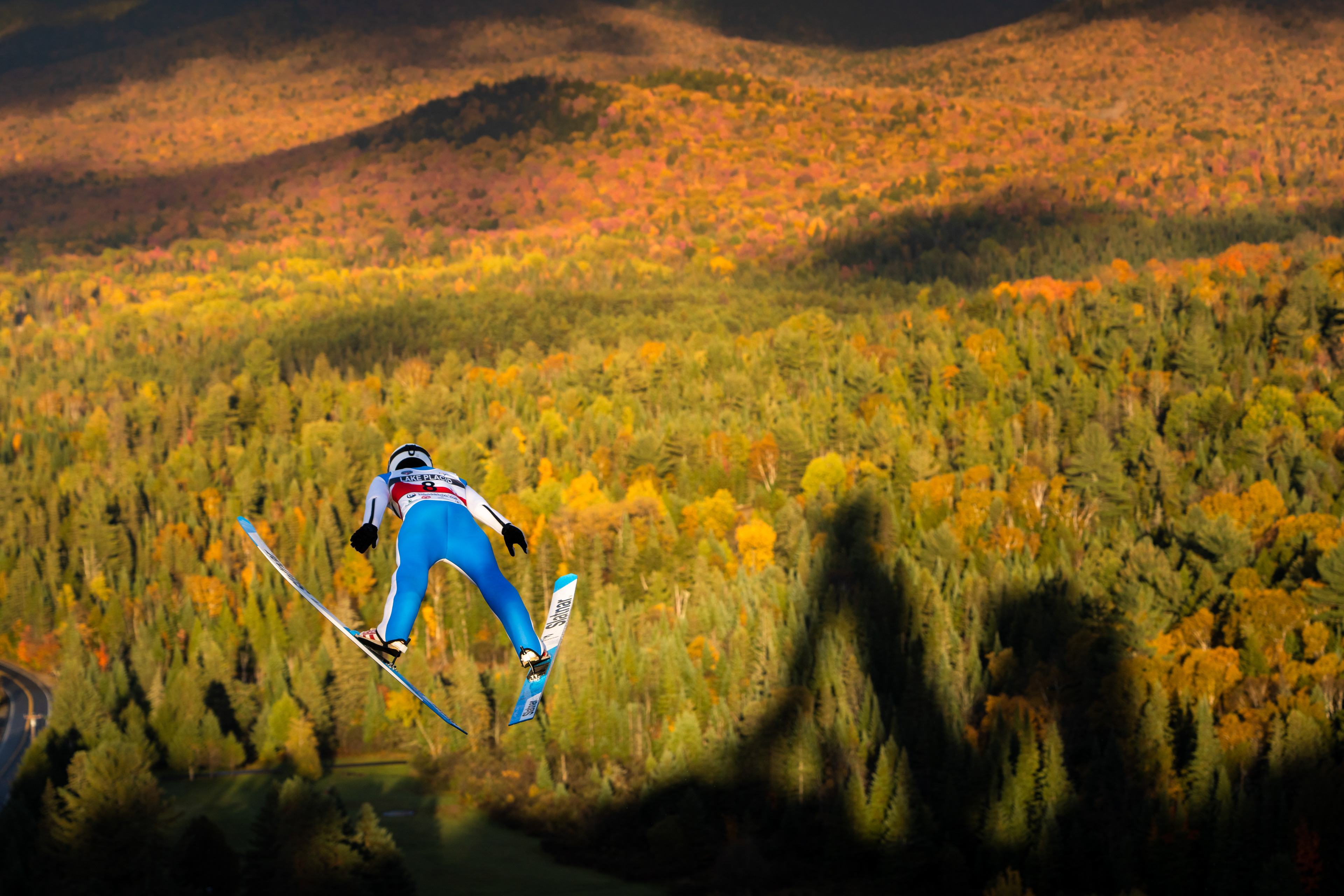 A male Ski jumper is flying across a sea of red, orange and yellow trees. The tiki jumping towers, off which he is flying, cast their shadows onto the treed background below! Leaf Peeping. Autumn. Fall leaves. Lake Placid, New York.