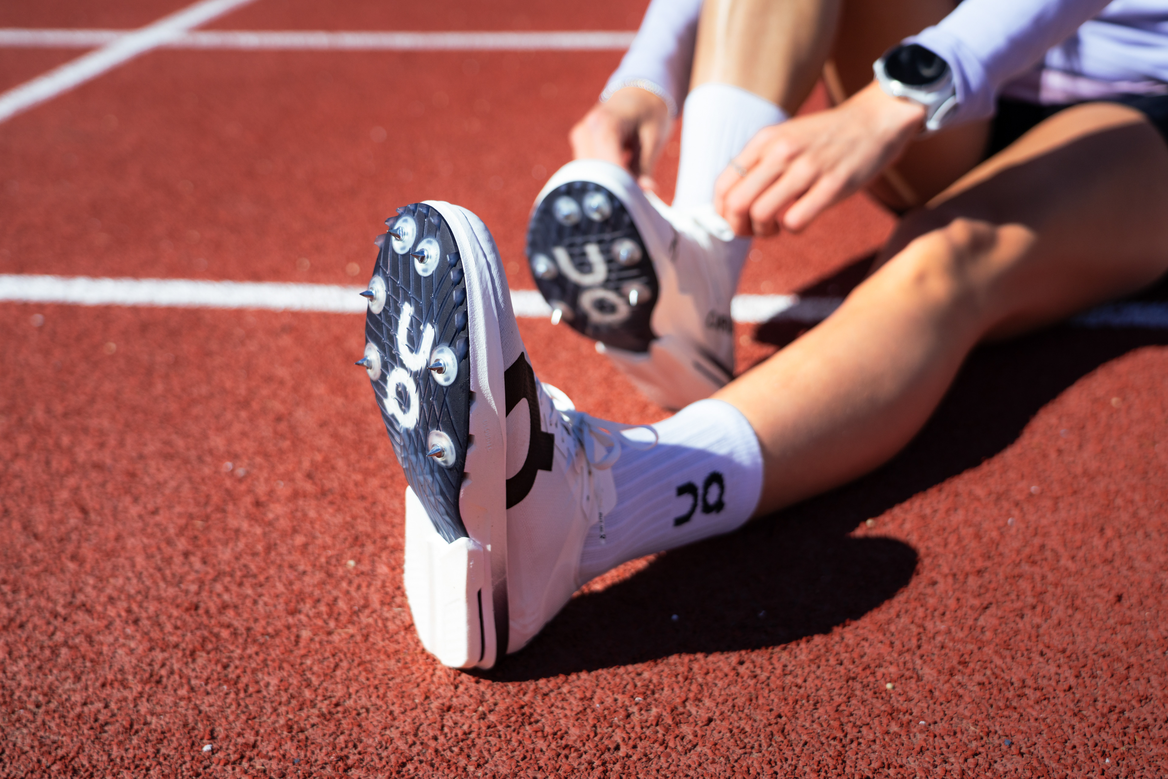 A runner ties her On Track Spikes while sitting on the red track surface.