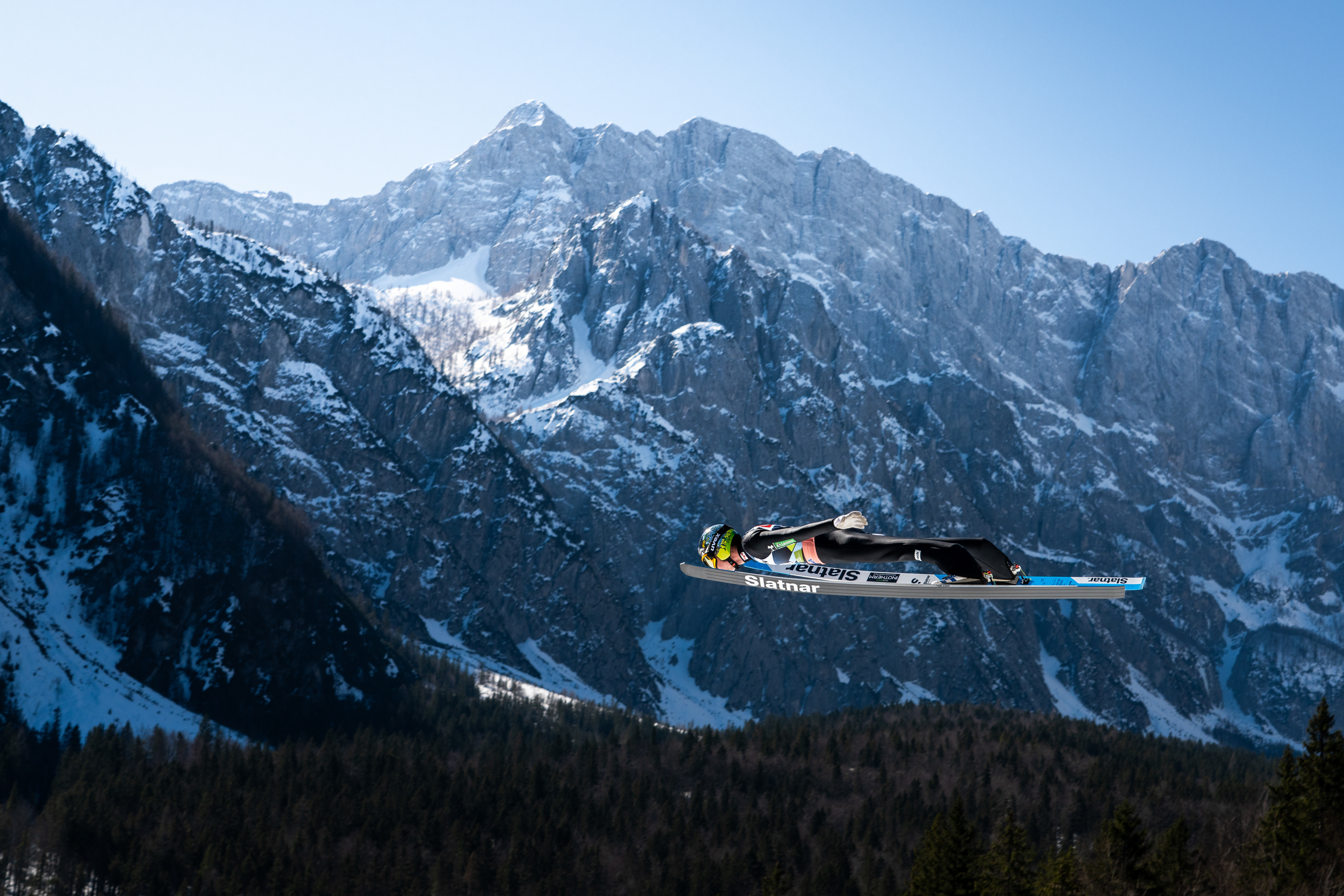 A male Ski Jumper with his face between the tips of his skis, flys infant of the dramatic Slovenian Mountain backdrop