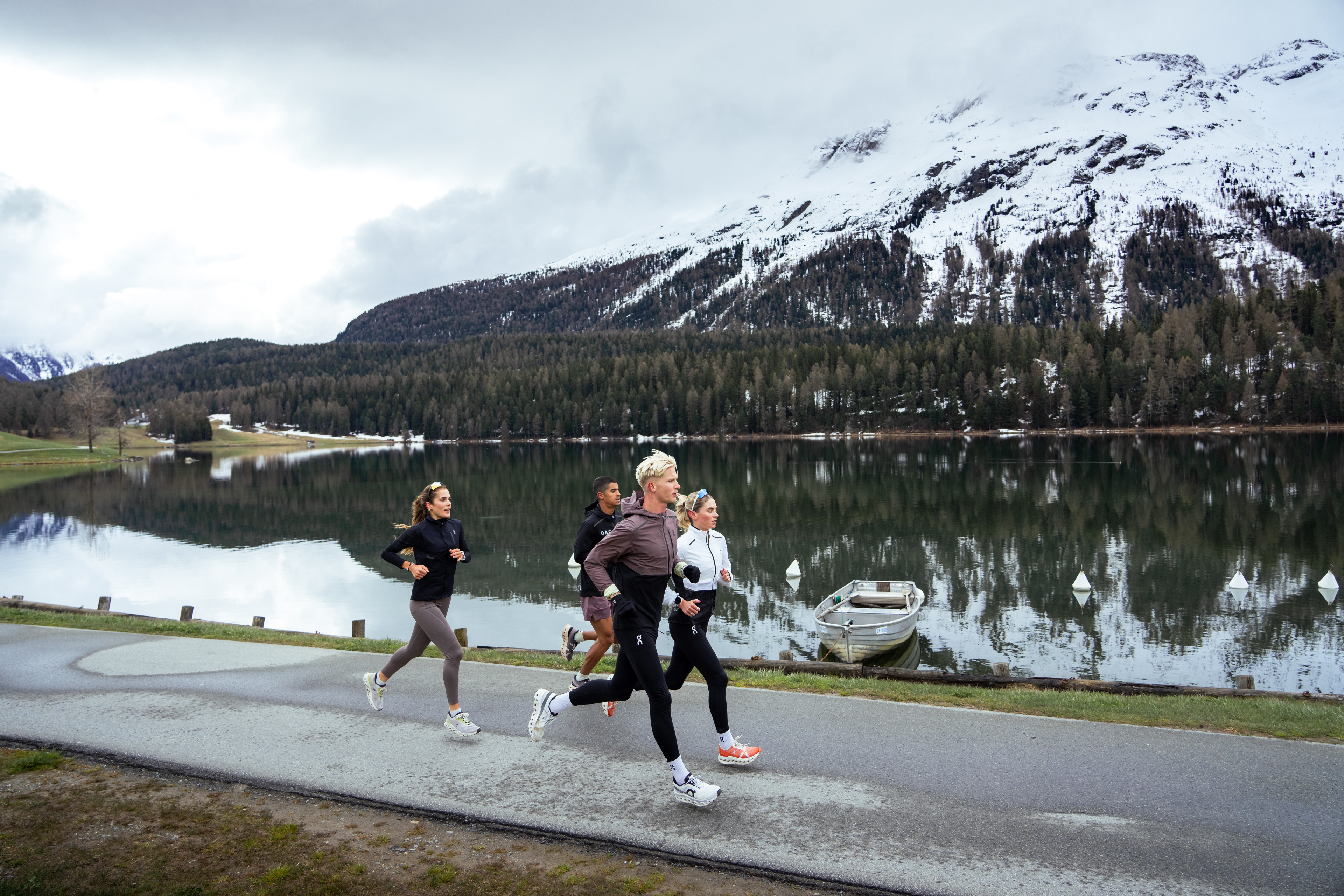 four athletes running on a paved path. There is a Lake, with a small boat and snow capped mountains in the background